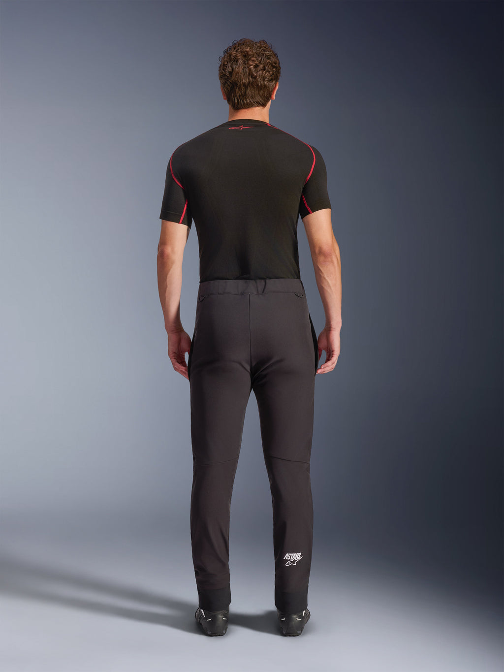 Alpinestars Aeroshell Hose, Crossover-Sport-Softshell-Motorradhose, Schwarz, Rückansicht kombiniert mit einem schwarzen kurzärmeligen Base-Layer, schmale Passform, vorgeformte Knie, weißes Astars-Logo am unteren linken Bein.