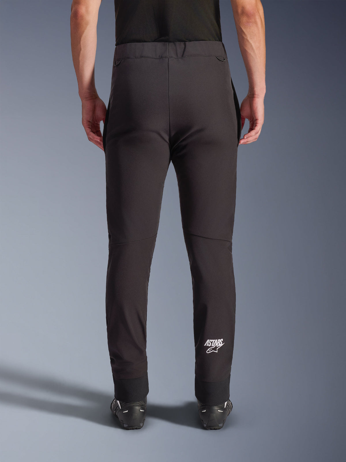 Alpinestars Aeroshell Hose, schwarze Softshell-Motorradhose, Rückansicht mit schmaler Passform und vorgeformten Knien, weißes Astars-Logo an der unteren rechten Wade, elastischer unterer Saum, Crossover-Design für Straßenfahrten