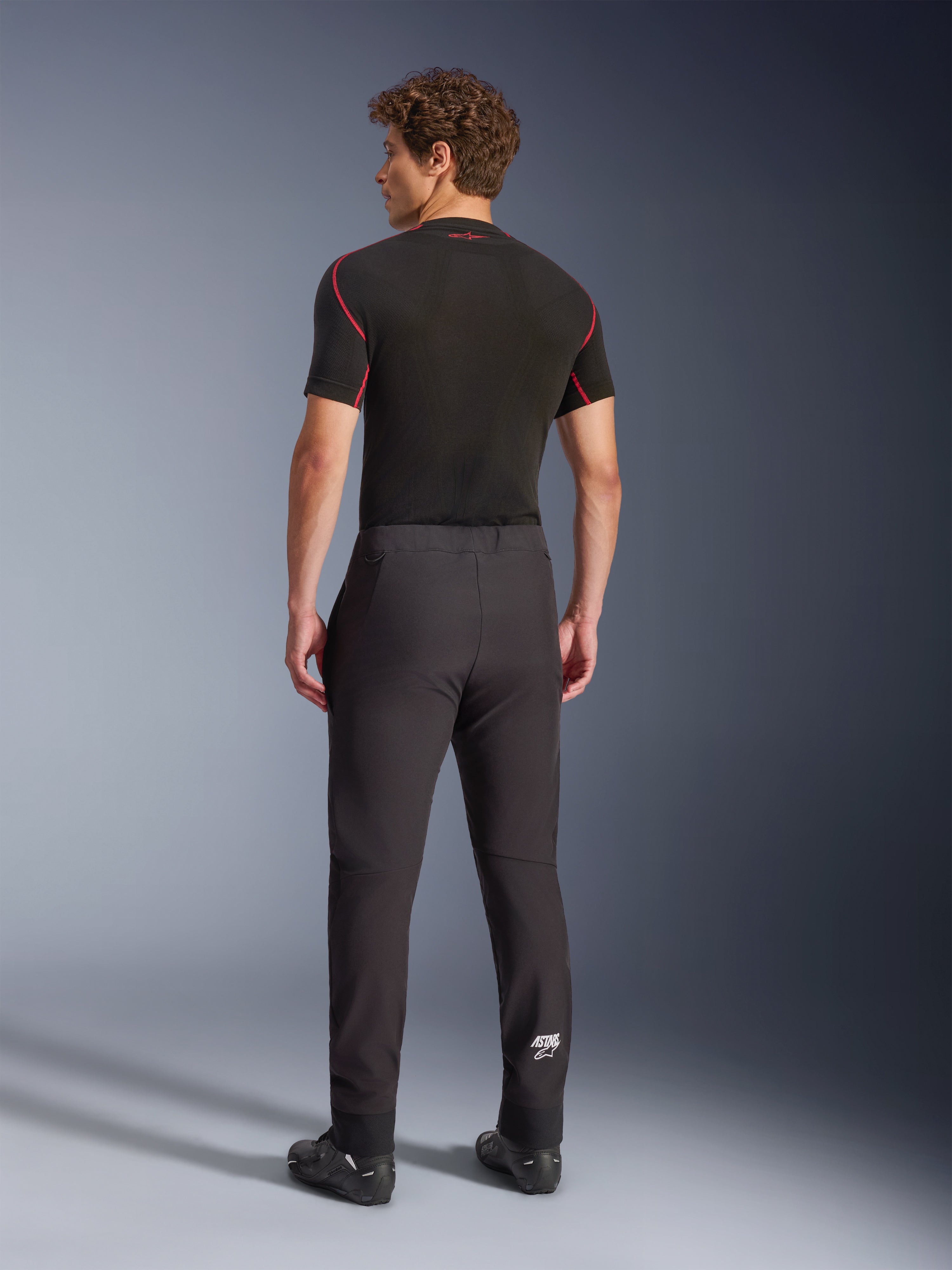 Alpinestars Aeroshell Hose, Schwarz, Softshell-Motorradhose, Rückansicht an einem Model, moderne schmal zulaufende Passform, weißes Astars-Logo am Unterschenkel, elastische Säume, kombiniert mit einem schwarzen kurzärmeligen Base Layer und Motorradschuhen.