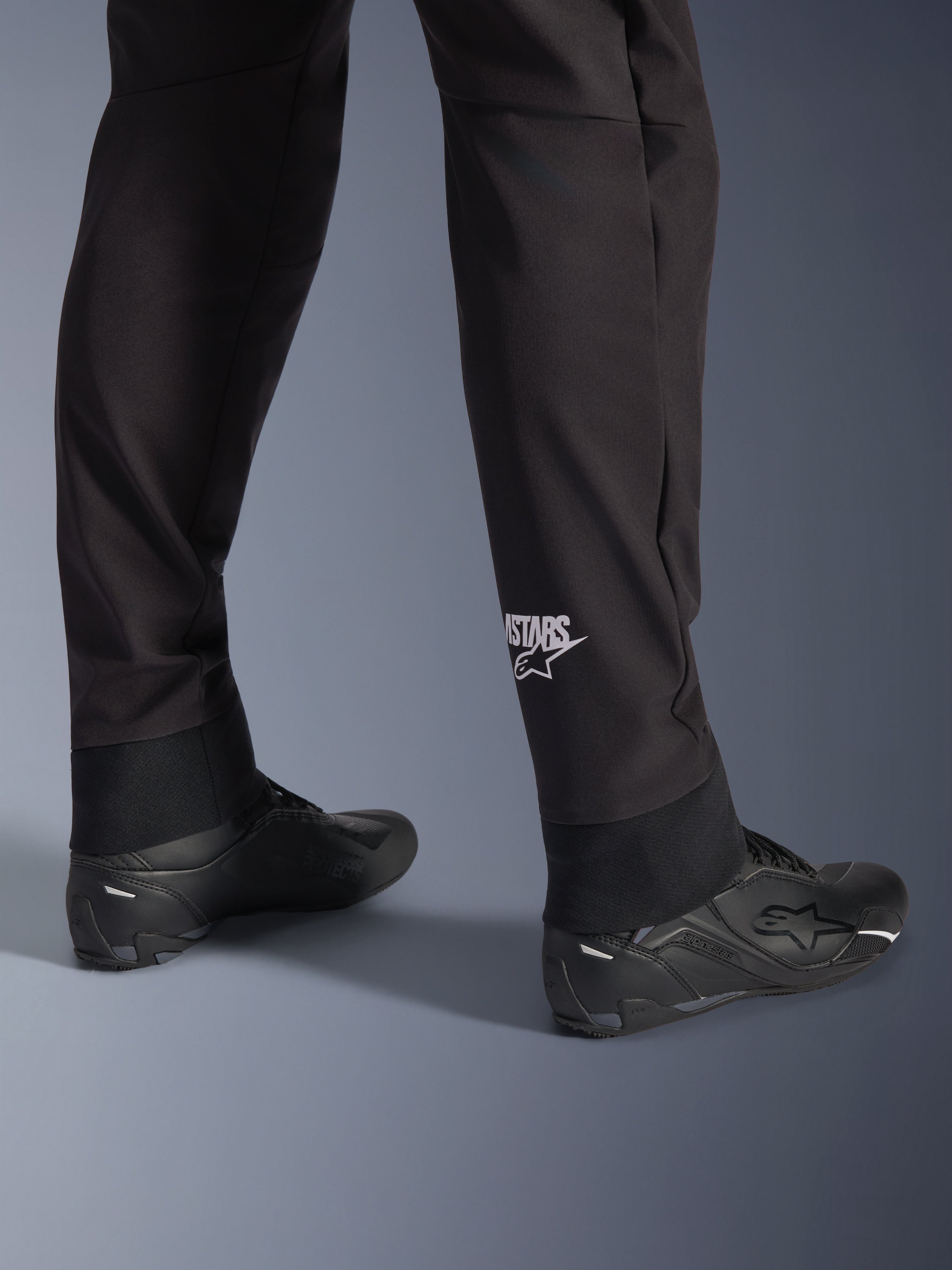 Alpinestars Aeroshell Hose, schwarze Softshell-Motorradhose, Nahaufnahme des Unterschenkels und des elastischen Saums, weißes Alpinestars-Logo an der Wade, kombiniert mit schwarzen Motorradschuhen, modernes Crossover-Sport-Design