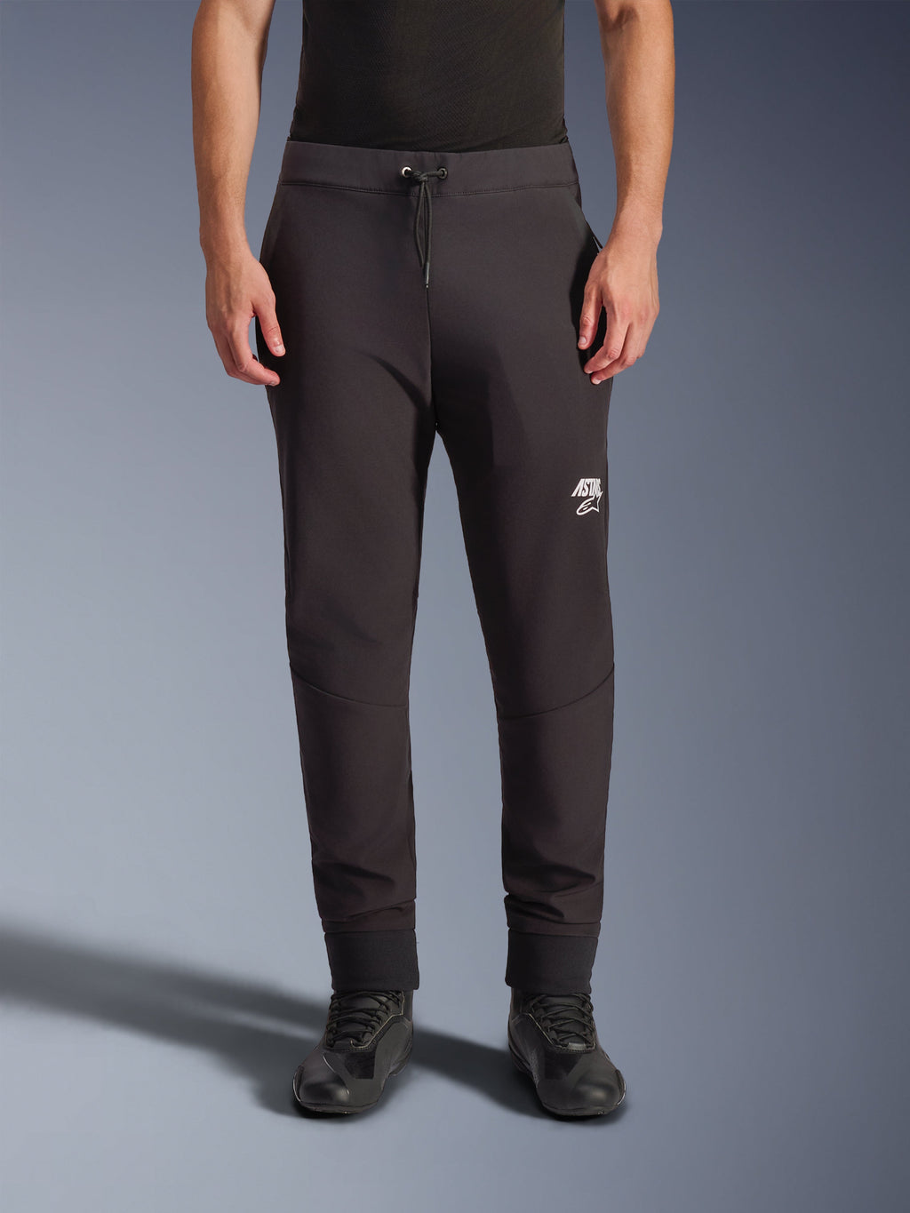 Alpinestars Aeroshell Hose, schwarze Softshell-Motorradhose, athletische Passform im Jogger-Stil mit Kordelzug und elastischen Bündchen, weißes Astars-Logo, getragen mit schwarzen Motorradschuhen, moderne Crossover-Ausrüstung für Straße und Freizeit