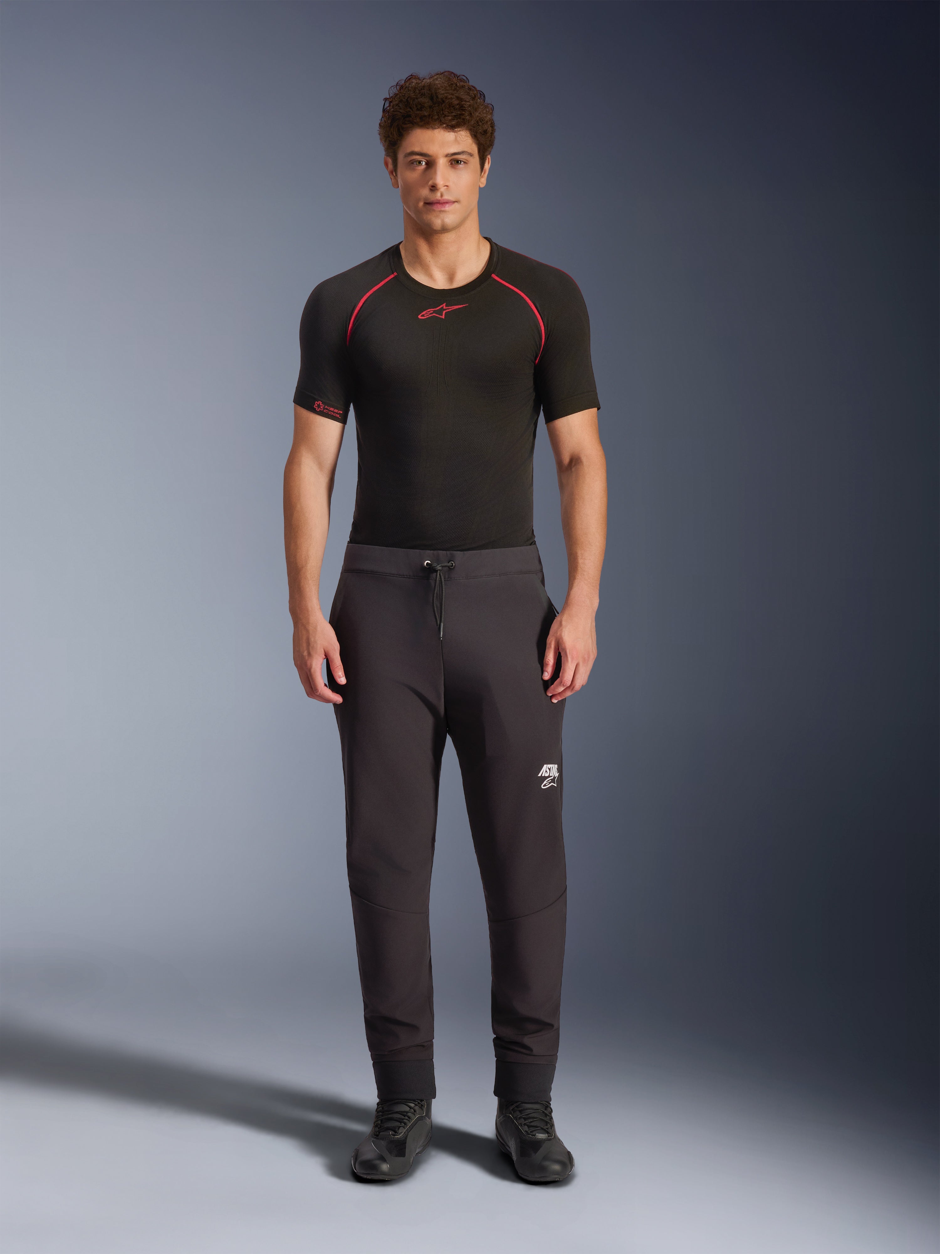Alpinestars Aeroshell Hose, schwarze Motorrad-Softshell-Hose, getragen mit einem schwarzen kurzärmeligen Base Layer und schwarzen Schuhen, mit elastischem Kordelzug am Bund, weißem Astars-Logo am Oberschenkel und elastischen Bündchen, moderne sportliche Silhouette für das Fahren auf der Straße