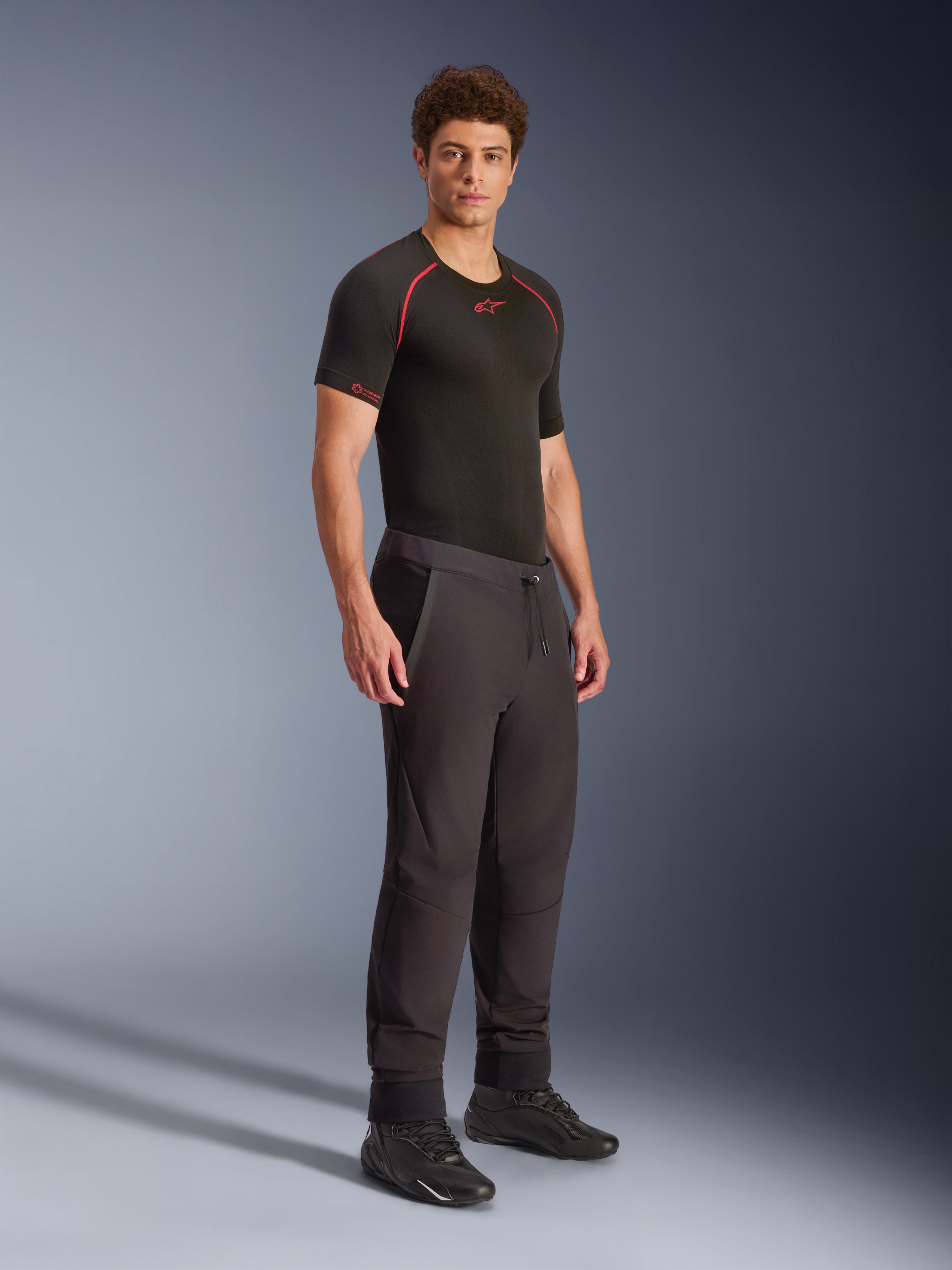 Alpinestars Aeroshell Hose, Schwarz, Crossover-Sport-Softshell-Motorradhose, getragen von einem männlichen Model mit einem schwarzen Performance-Base-Layer und schwarzen Schuhen, mit Kordelzug am Bund und elastischen Säumen.