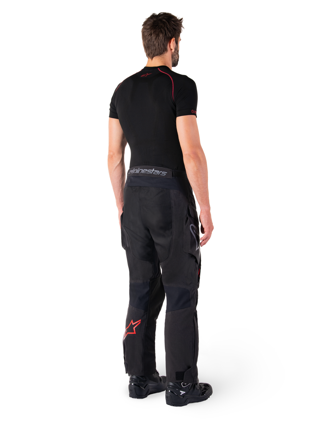 Alpinestars Hyde XT Drystar® Hose, Adventure-Tourenhose, Schwarz und Hellrot, Rückansicht eines Fahrers mit schwarzem Base-Layer und schwarzen Stiefeln, strapazierfähige Textilkonstruktion mit rotem Stern-Logo am Unterschenkel und grauem Alpinestars-Branding am Bund