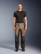 Männliches Model in Alpinestars Andes V4 Drystar® Hose in Walnut Black, Adventure-Motorradhose kombiniert mit einem schwarzen Base Layer und Stiefeln, mit orangefarbenen Reißverschluss-Akzenten an den Oberschenkeln und verstärkten schwarzen Einsätzen für Strapazierfähigkeit in jedem Gelände.