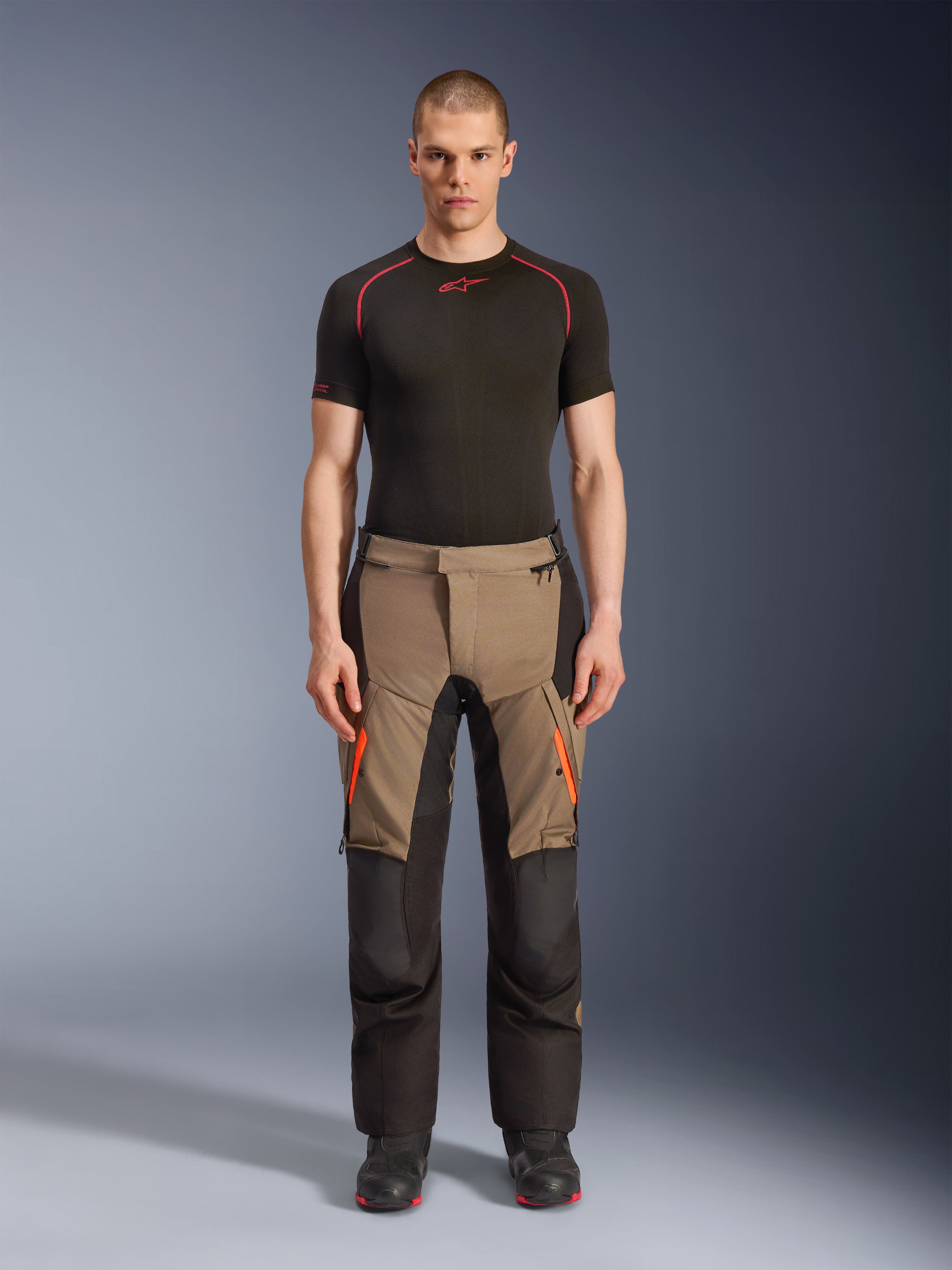 Männliches Model in Alpinestars Andes V4 Drystar® Hose in Walnut Black, Adventure-Motorradhose kombiniert mit einem schwarzen Base Layer und Stiefeln, mit orangefarbenen Reißverschluss-Akzenten an den Oberschenkeln und verstärkten schwarzen Einsätzen für Strapazierfähigkeit in jedem Gelände.