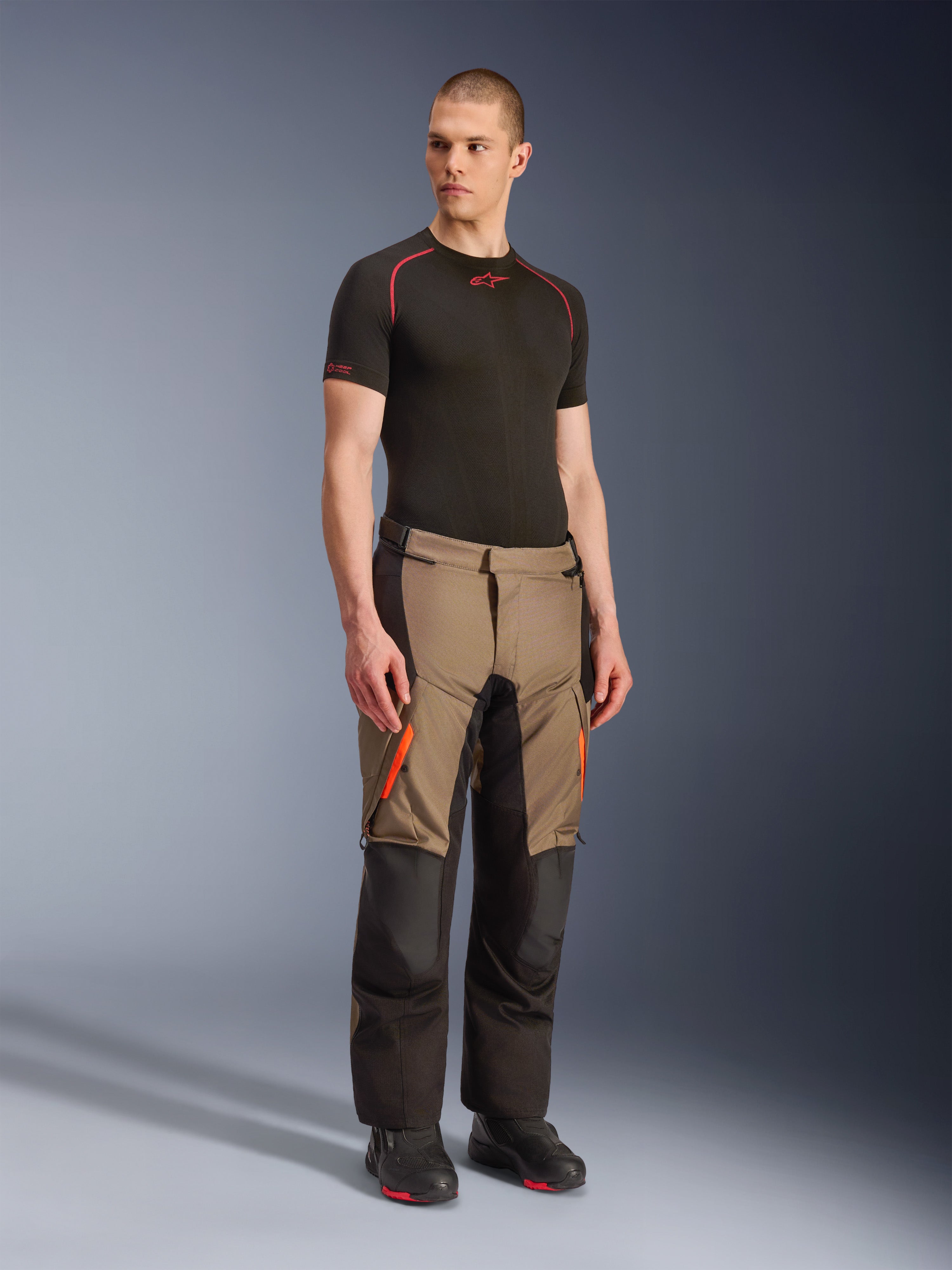 Alpinestars Andes V4 Drystar® Hose, Motorrad-Adventure-Touring-Hose, Walnut Black mit orangefarbenen Akzenten, getragen von einem Model mit schwarzem Kurzarm-Baselayer und schwarzen Stiefeln, verstärkte Textileinsätze und Cargotaschen, robustes Design für Fahrten bei jedem Wetter