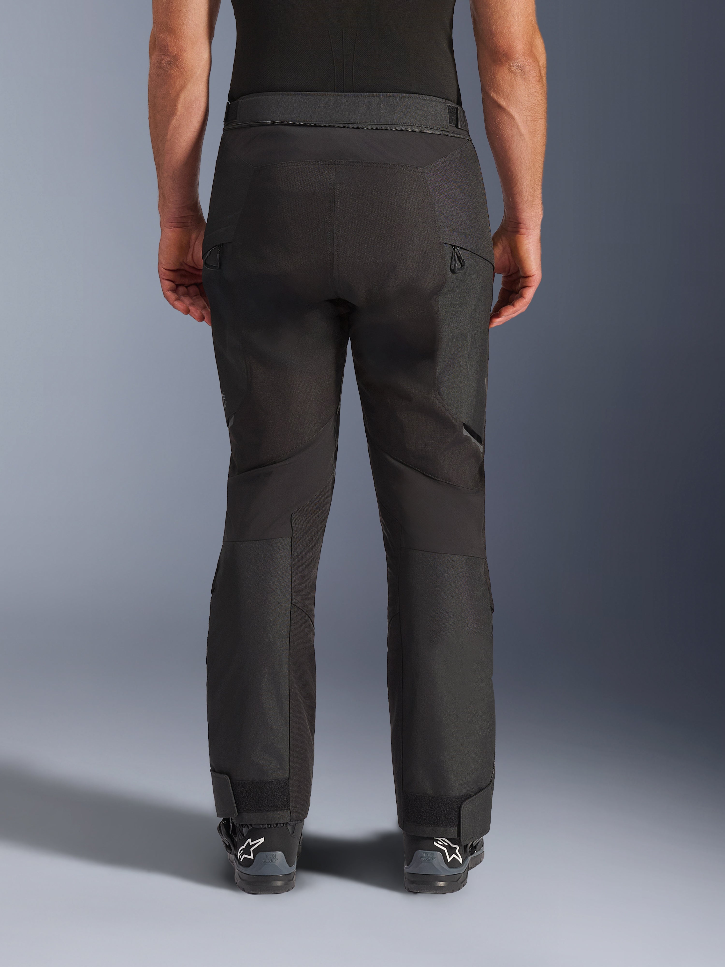 Alpinestars Cusco Drystar® Hose, Motorradhose, Schwarz, Rückansicht getragen von einem Fahrer, mit strukturierten Textilpaneelen, Belüftungsöffnungen mit Reißverschluss an den Oberschenkeln und verstellbaren Knöchelverschlüssen, kombiniert mit schwarzen Alpinestars-Stiefeln.