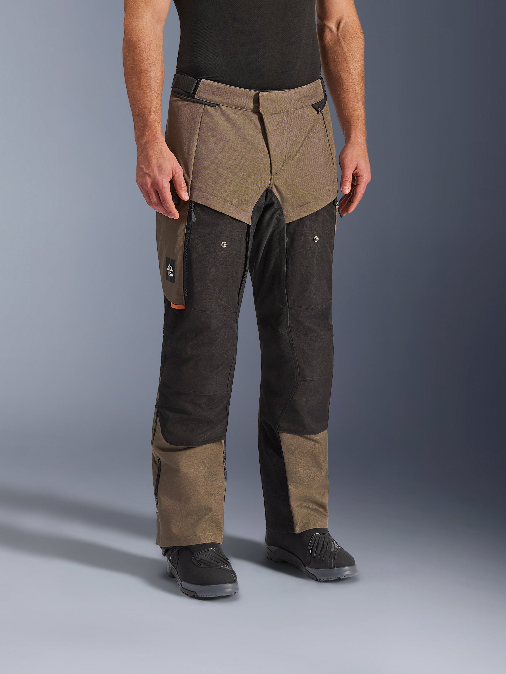 Alpinestars Cusco Drystar® Hose, Adventure-Motorradhose, Walnut Black, abgebildet an einem Modell mit schwarzen Stiefeln, in einem braun-schwarzen Multi-Panel-Design mit Reißverschlusstaschen und verstärkten Knien für Touren