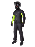 Alpinestars Bogota' Pro Drystar® 3-Jahreszeiten-Hose, Schwarz, Fahrer in kompletter Adventure-Touring-Ausrüstung inklusive schwarzer Jacke mit neongelben Akzenten, schwarzem Helm, Handschuhen und Stiefeln, zeigt die wasserdichte Drystar®-Membran, die über der Hose getragen wird
