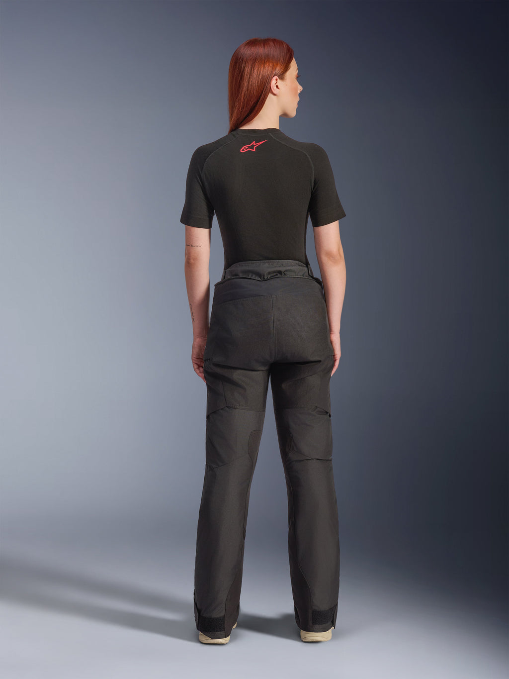 Rückansicht eines Models in der Alpinestars Stella Andes V4 Drystar® Hose in Schwarz, kombiniert mit einem schwarzen, kurzärmeligen Baselayer mit rotem Alpinestars-Logo, Adventure-Touring-Motorradausrüstung mit strapazierfähiger Textilkonstruktion.