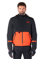 Alpinestars Mootant Waterproof Parka, modulare Motorradjacke in Spicy Orange und Schwarz, von einem Model mit passenden Handschuhen getragen, Kapuzendesign mit zweifarbigen Einsätzen, Frontreißverschluss und verstellbare Bündchen, moderne urbane Motorradbekleidung