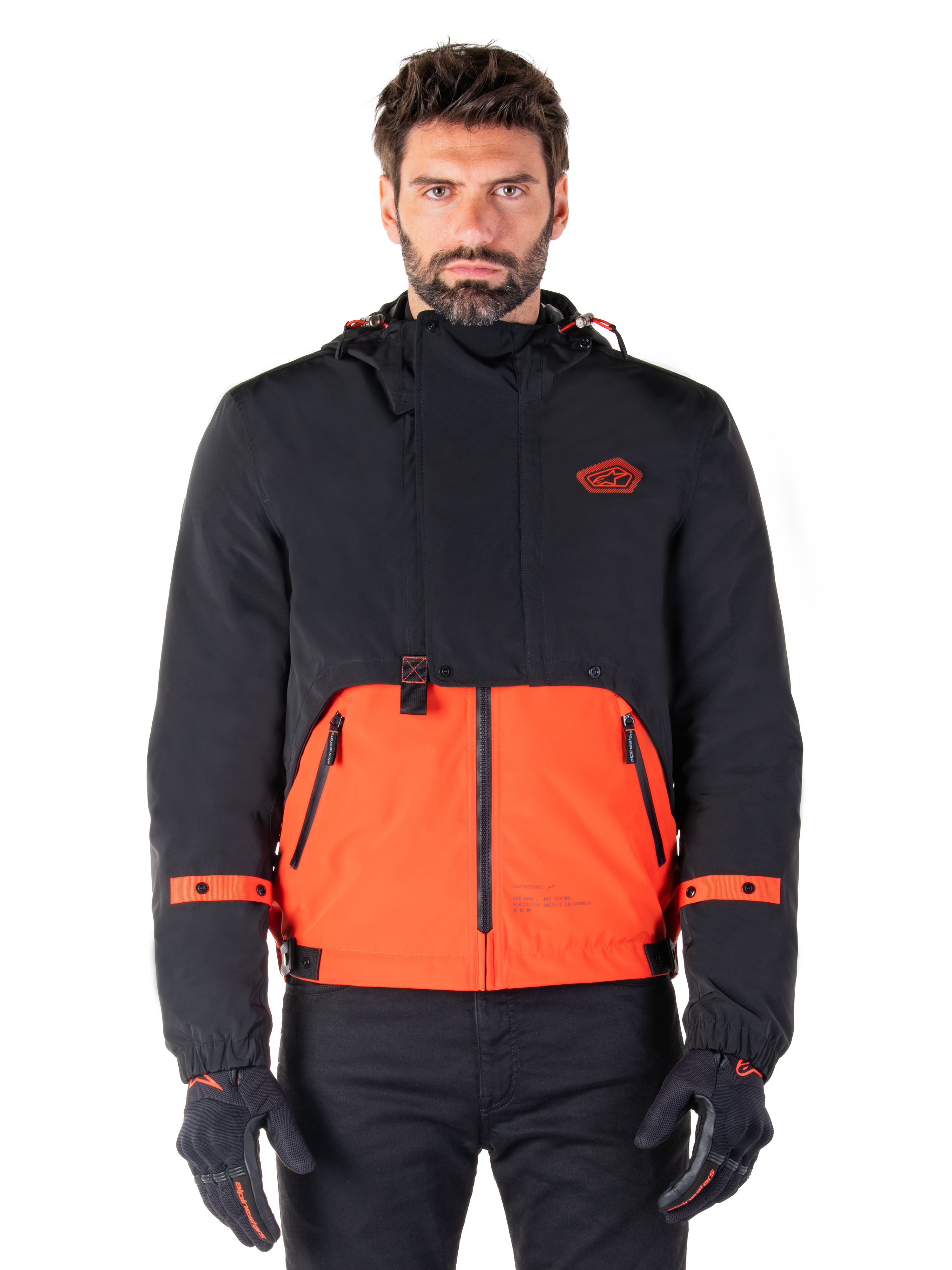 Alpinestars Mootant Waterproof Parka, modulare Motorradjacke in Spicy Orange und Schwarz, von einem Model mit passenden Handschuhen getragen, Kapuzendesign mit zweifarbigen Einsätzen, Frontreißverschluss und verstellbare Bündchen, moderne urbane Motorradbekleidung