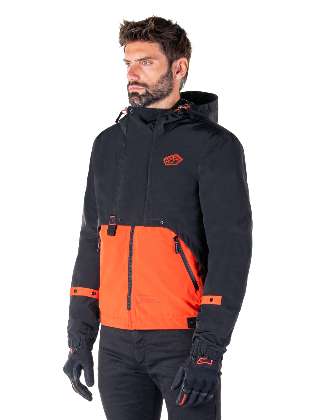 Mann im Alpinestars Mootant Wasserdichten Parka in Spicy Orange und Schwarz, eine modulare Motorradjacke mit einer schwarzen Kapuzen-Oberschicht und einem leuchtend orangefarbenen Unterteil, kombiniert mit schwarzen Motorradhandschuhen und -hosen.