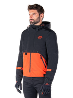 Mann im Alpinestars Mootant Wasserdichten Parka in Spicy Orange und Schwarz, eine modulare Motorradjacke mit einer schwarzen Kapuzen-Oberschicht und einem leuchtend orangefarbenen Unterteil, kombiniert mit schwarzen Motorradhandschuhen und -hosen.