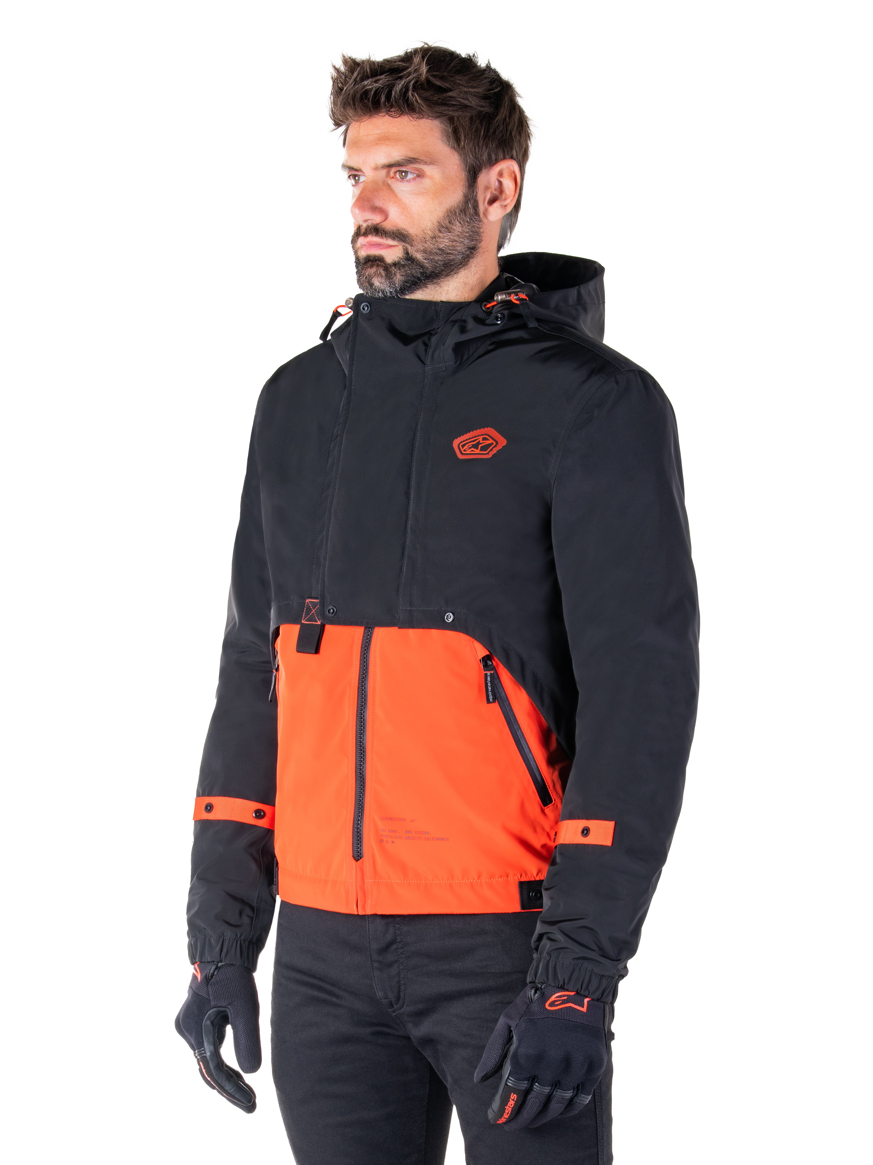 Mann im Alpinestars Mootant Wasserdichten Parka in Spicy Orange und Schwarz, eine modulare Motorradjacke mit einer schwarzen Kapuzen-Oberschicht und einem leuchtend orangefarbenen Unterteil, kombiniert mit schwarzen Motorradhandschuhen und -hosen.