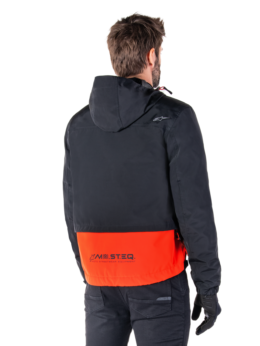 Alpinestars Mootant Wasserdichter Parka, Spicy Orange und Schwarz, Rückansicht an einem männlichen Model, verfügt über einen schwarzen Oberteil mit Kapuze und einen leuchtend orangen unteren Bereich mit MO.ST.EQ. Branding, urbane zeitgemäße Motorradjacke, getragen mit schwarzer Hose und Handschuhen