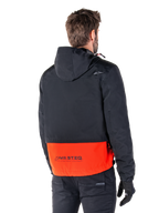 Alpinestars Mootant Wasserdichter Parka, Spicy Orange und Schwarz, Rückansicht an einem männlichen Model, verfügt über einen schwarzen Oberteil mit Kapuze und einen leuchtend orangen unteren Bereich mit MO.ST.EQ. Branding, urbane zeitgemäße Motorradjacke, getragen mit schwarzer Hose und Handschuhen