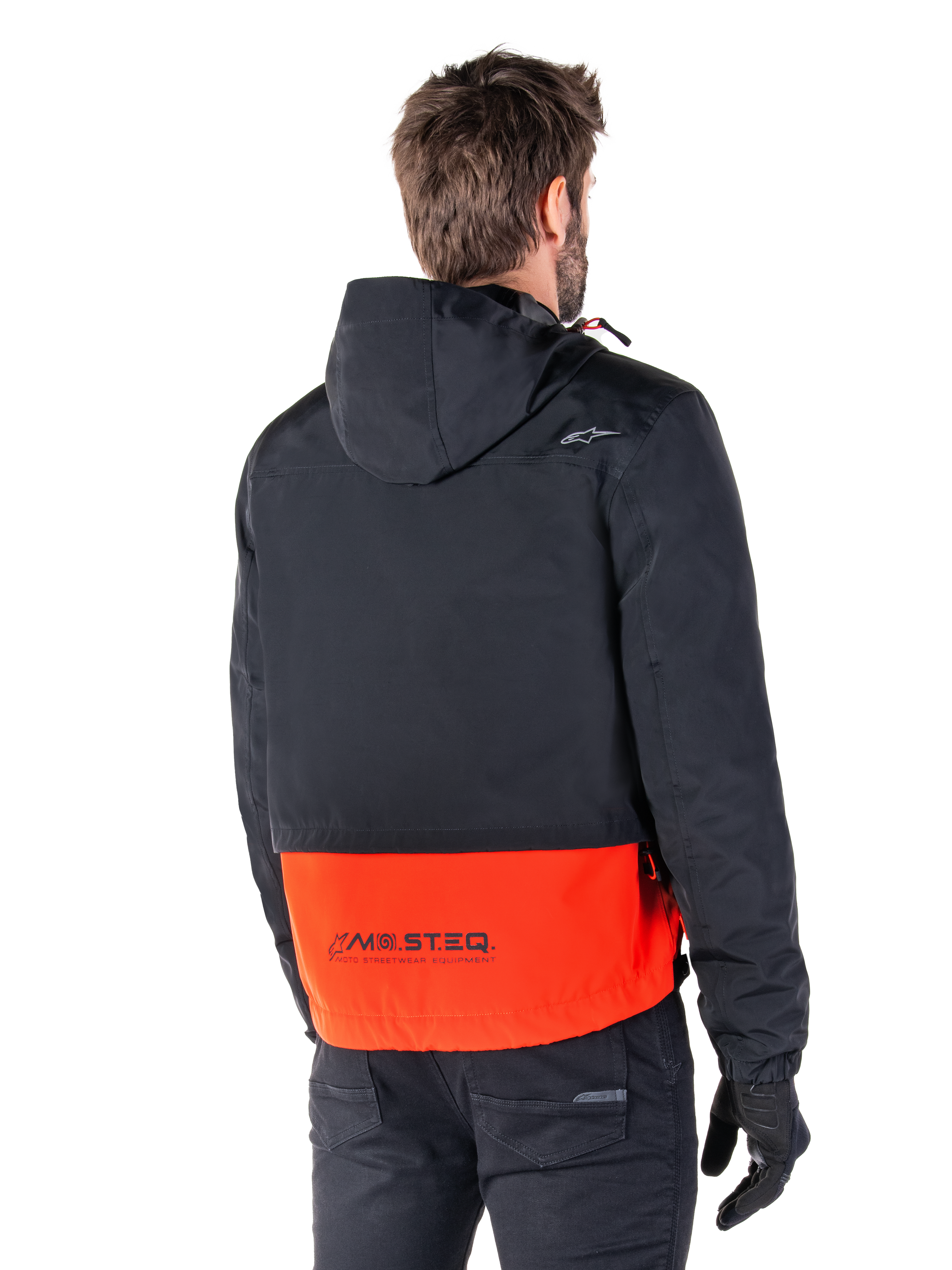 Alpinestars Mootant Wasserdichter Parka, Spicy Orange und Schwarz, Rückansicht an einem männlichen Model, verfügt über einen schwarzen Oberteil mit Kapuze und einen leuchtend orangen unteren Bereich mit MO.ST.EQ. Branding, urbane zeitgemäße Motorradjacke, getragen mit schwarzer Hose und Handschuhen