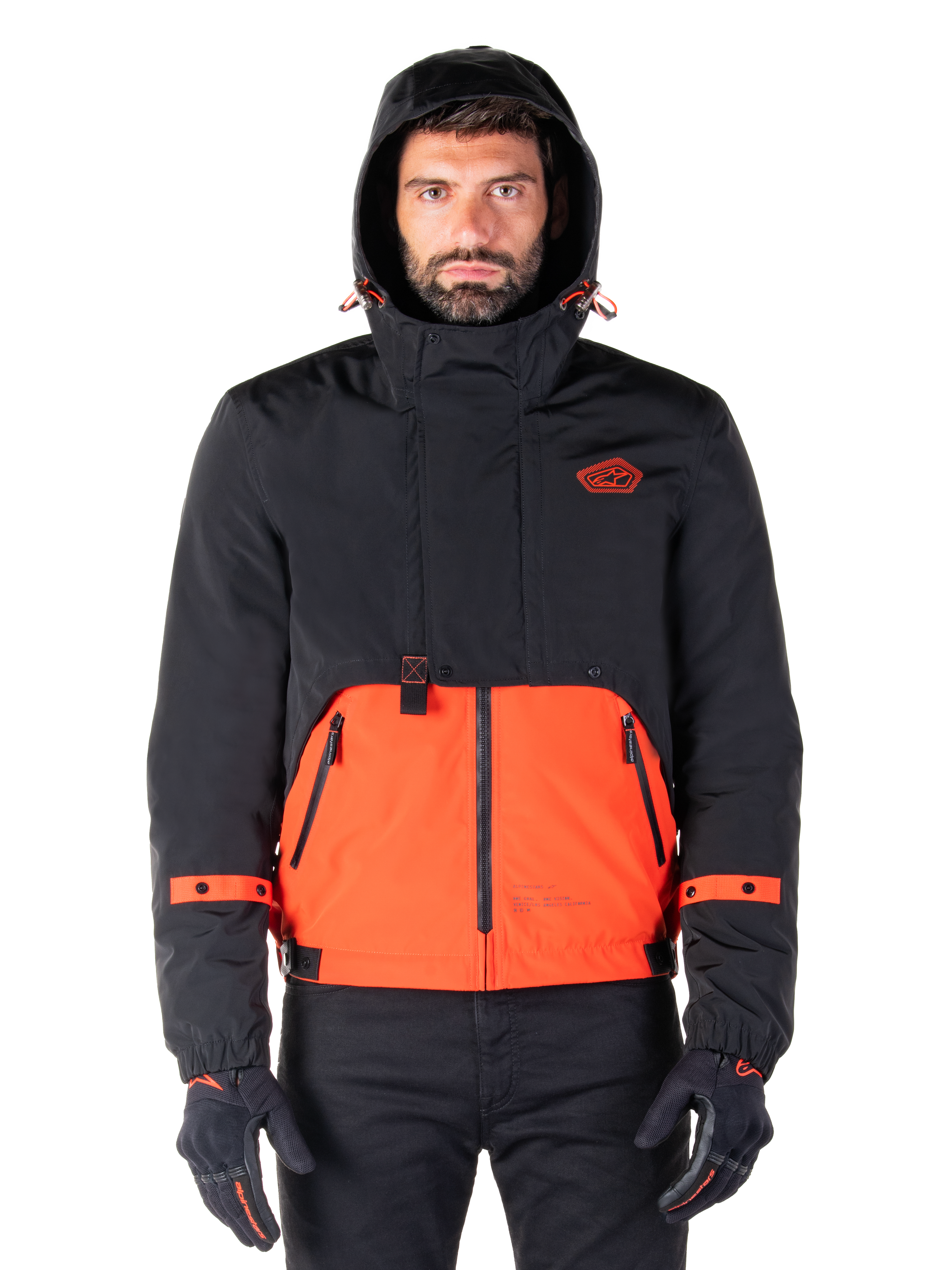 Alpinestars Mootant wasserdichter Parka, Motorradjacke, Spicy Orange und Schwarz, modulares Allwetter-Design mit Kapuze, Reißverschlusstaschen vorne und orangefarbene Riemendetails an den Ärmeln, getragen von einem Model mit schwarzen Handschuhen und Hosen