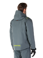 Alpinestars Morush Wasserdichter Parka in Urban Gray, Rückansicht, mit gelbem MO.ST.EQ. Branding am unteren Rücken und an der Schulter, minimalistisches Kapuzendesign, getragen mit passender grauer Motorradhose und schwarzen Handschuhen, urbane Motorrad-Streetwear.