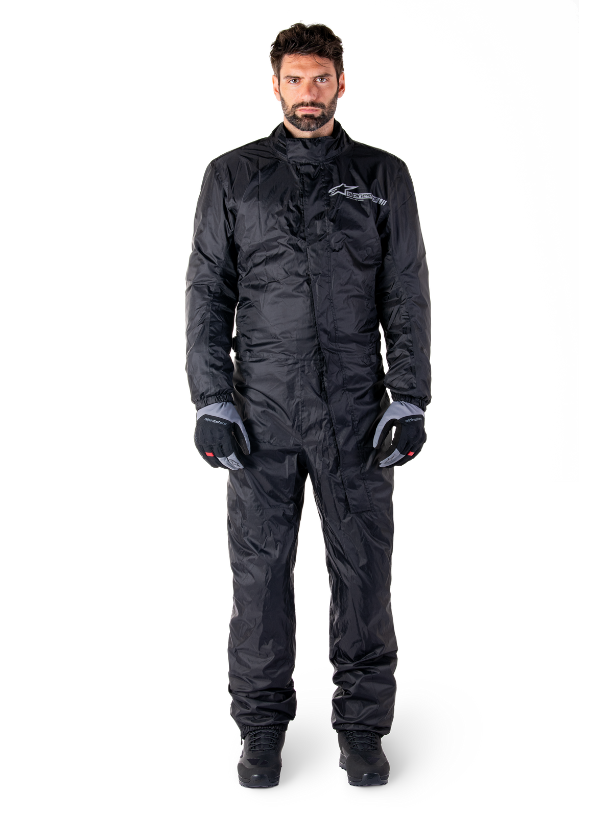 Alpinestars Hurricane Rain V2 Anzug, Ganzkörper-Motorrad-Regenanzug in Schwarz, wasserdichte Schutzausrüstung mit hohem Kragen und Front-Sturmverschluss, weißes Logo auf der Brust, getragen mit schwarz-grauen Handschuhen und Stiefeln