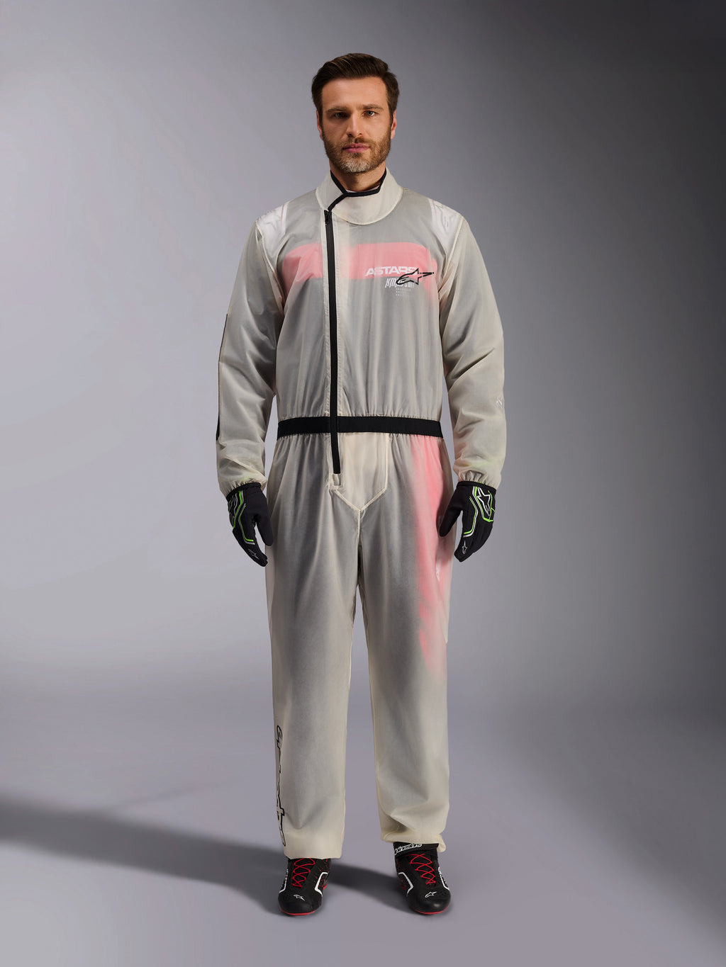 Alpinestars Kart Regenanzug, Transparent, transparenter wasserdichter Überanzug getragen von einem Model, mit schwarzem asymmetrischem Reißverschluss und Brust-Logo, darunter sichtbarer rot-weißer Rennanzug, kombiniert mit schwarz-neongrünen Handschuhen und schwarzen Rennstiefeln.