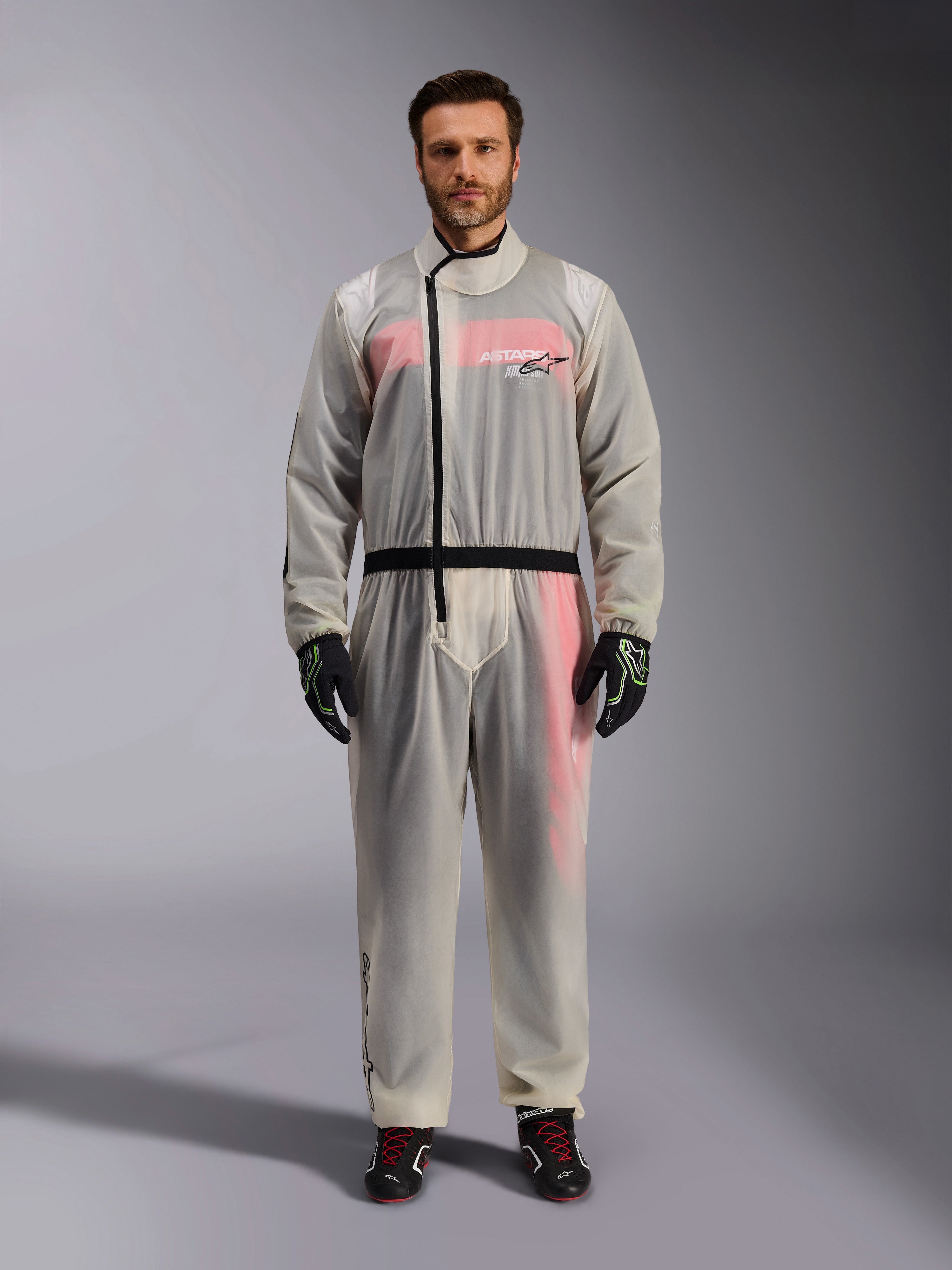 Alpinestars Kart Regenanzug, Transparent, transparenter wasserdichter Überanzug getragen von einem Model, mit schwarzem asymmetrischem Reißverschluss und Brust-Logo, darunter sichtbarer rot-weißer Rennanzug, kombiniert mit schwarz-neongrünen Handschuhen und schwarzen Rennstiefeln.
