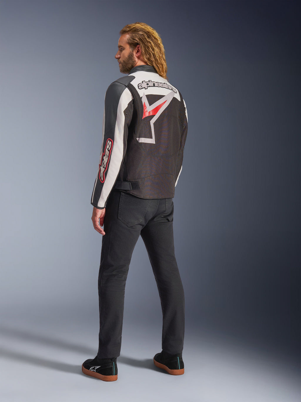Alpinestars Axton Jacket, Motorcycle Jacket, Black Off White and Bright Red, rear view worn by male model, featuring large Alpinestars star logo and wordmark across the back, vintage racing-inspired design with accordion panels, paired with black pants and sneakers