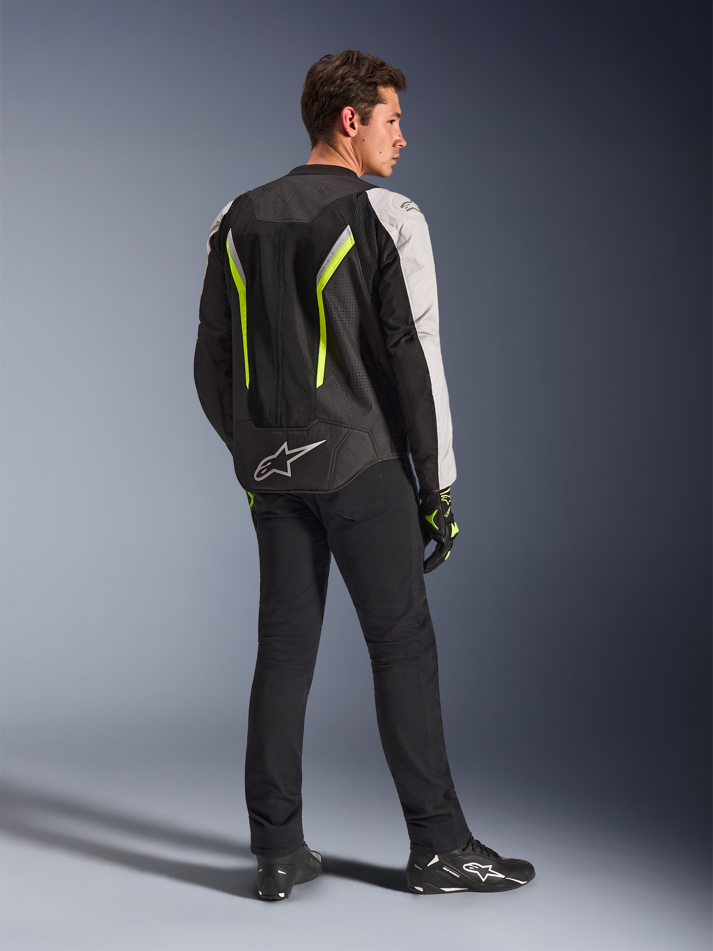 Alpinestars AST V3 Air Jacket in Black Silver Yellow Fluo, textile motorcycle jacket worn by male rider from behind, featuring extensive mesh panels and fluorescent yellow accents, paired with black riding pants, black gloves, and black Alpinestars shoes, showcasing technical gear for sport riding.