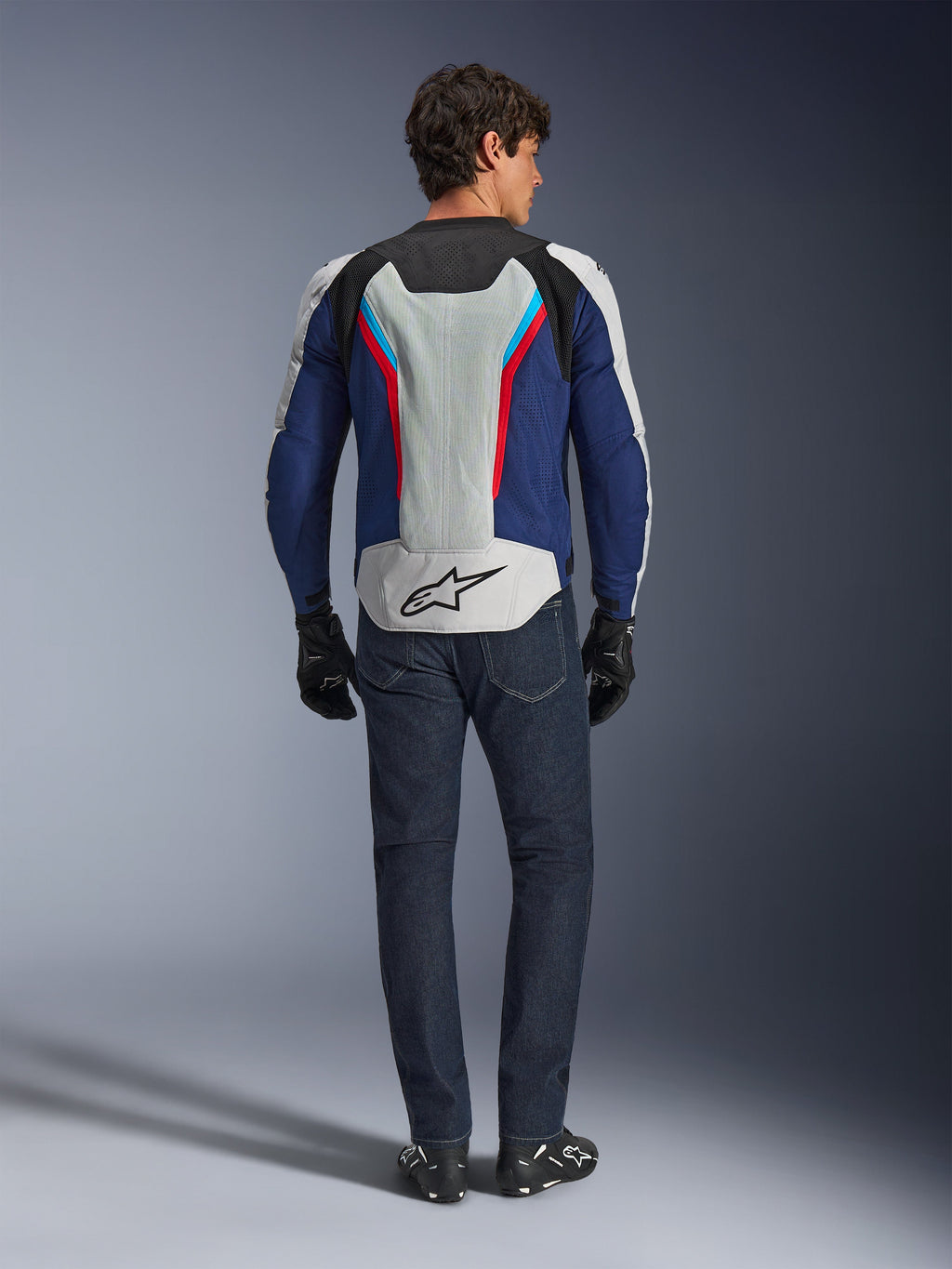 Alpinestars AST V3 Air Jacke, Rückansicht, Silber, Tiefblau und Hellrot, mit einem großen Mesh-Belüftungseinsatz auf dem Rücken und Alpinestars-Logo, getragen mit dunkelblauer Jeans und schwarzen Handschuhen, sportliche Motorradbekleidung für warmes Wetter