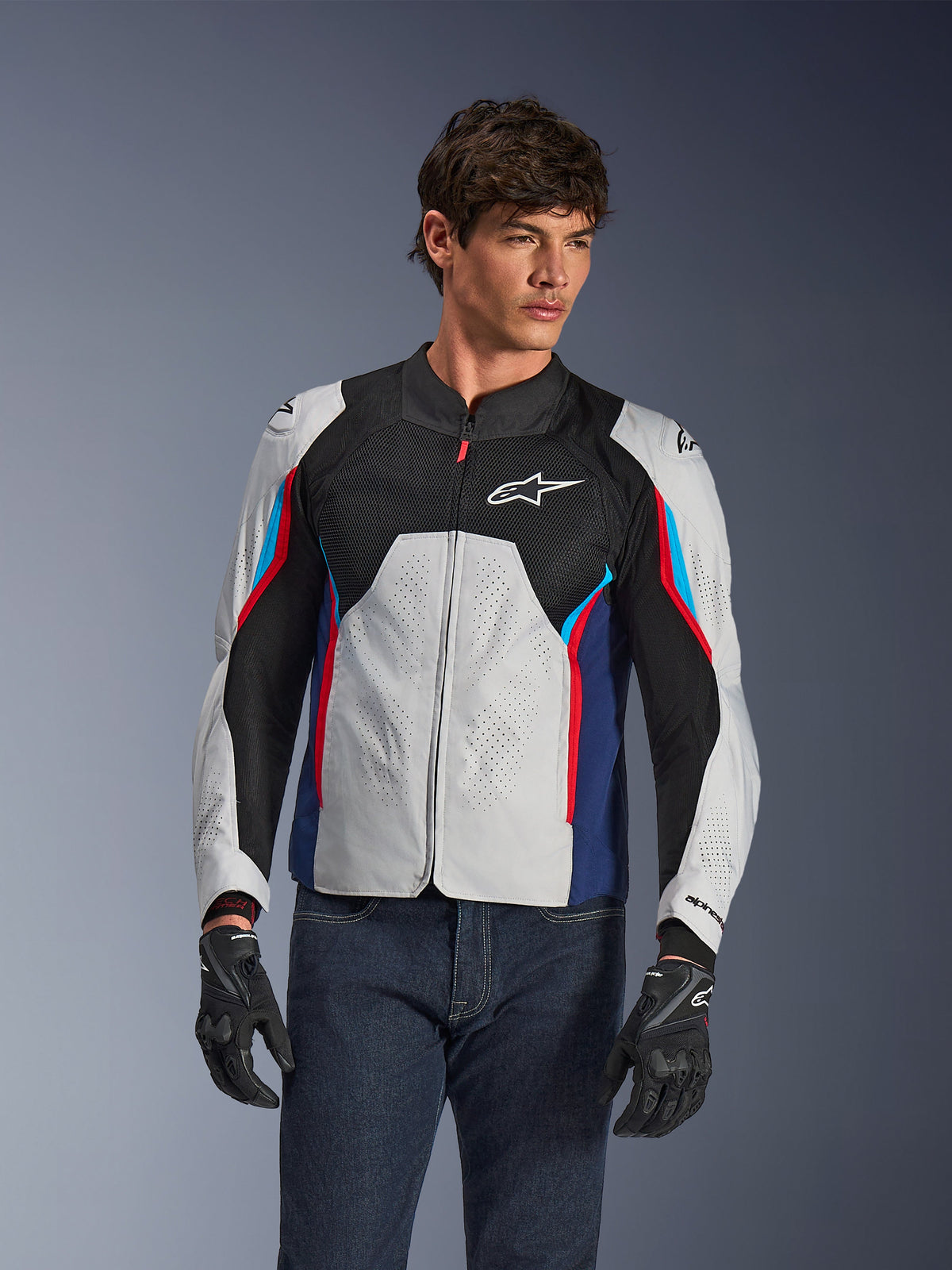 Alpinestars AST V3 Air Jacket in Silver Deep Blue Bright Red, worn by a model with dark jeans and black gloves, featuring perforated silver panels, black mesh ventilation, and sporty red and blue accents.
