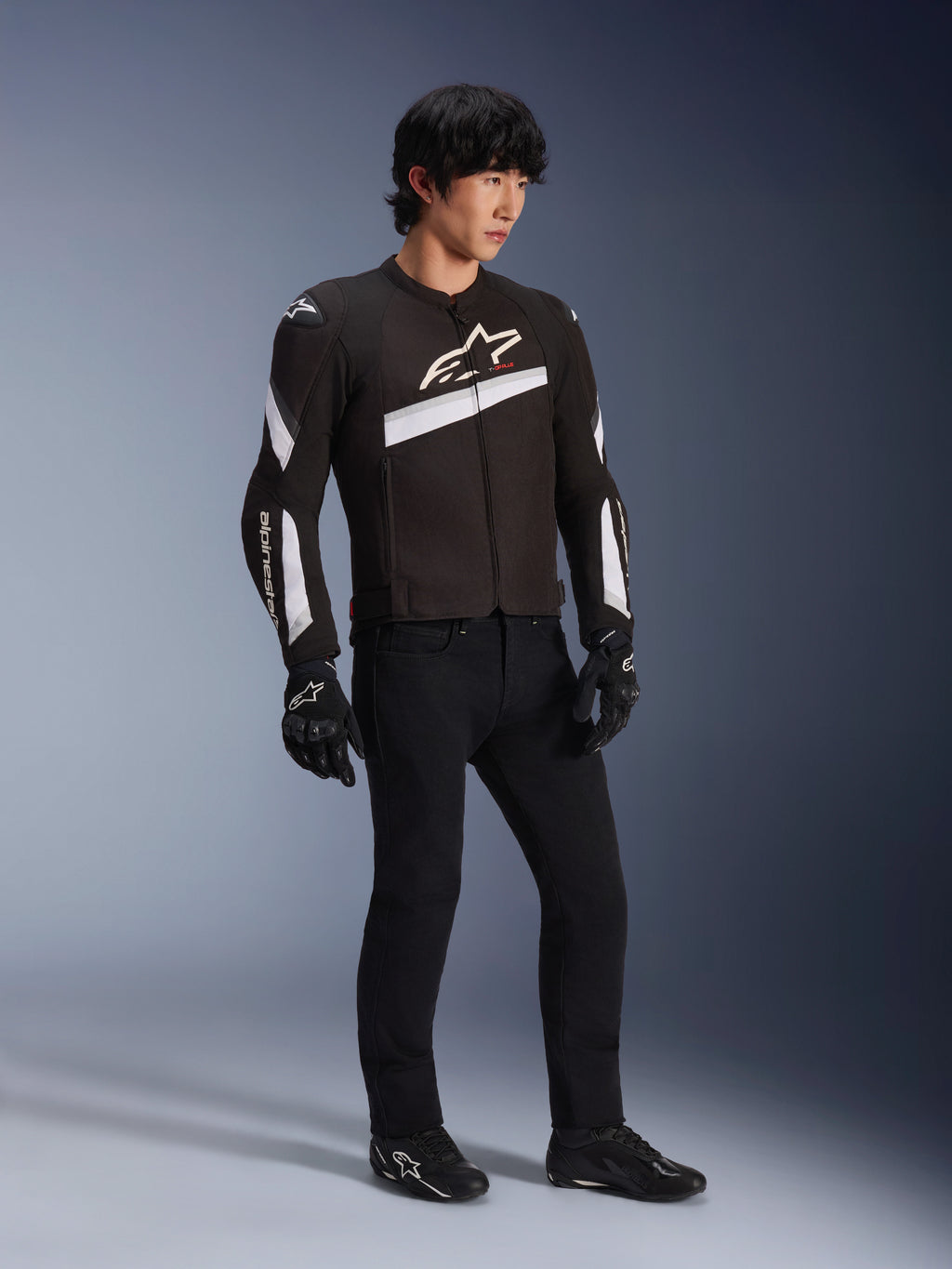 Alpinestars T-GP Plus R V4 Jacke, schwarz-weiße Textil-Motorradjacke, getragen von einem Model mit schwarzer Motorradhose und Handschuhen, mit weißen Alpinestars-Logos auf Brust und Ärmeln, sportliches Design für Performance auf der Straße