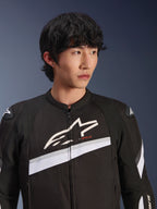 Alpinestars T-GP Plus R V4 Jacke, Schwarz und Weiß, Textil-Motorradjacke getragen von einem männlichen Model, großes weißes Stern-Logo auf der Brust, weiße diagonale Streifen, DFS Lite Schulterprotektoren, sportlich inspirierte Street-Riding-Ausrüstung.