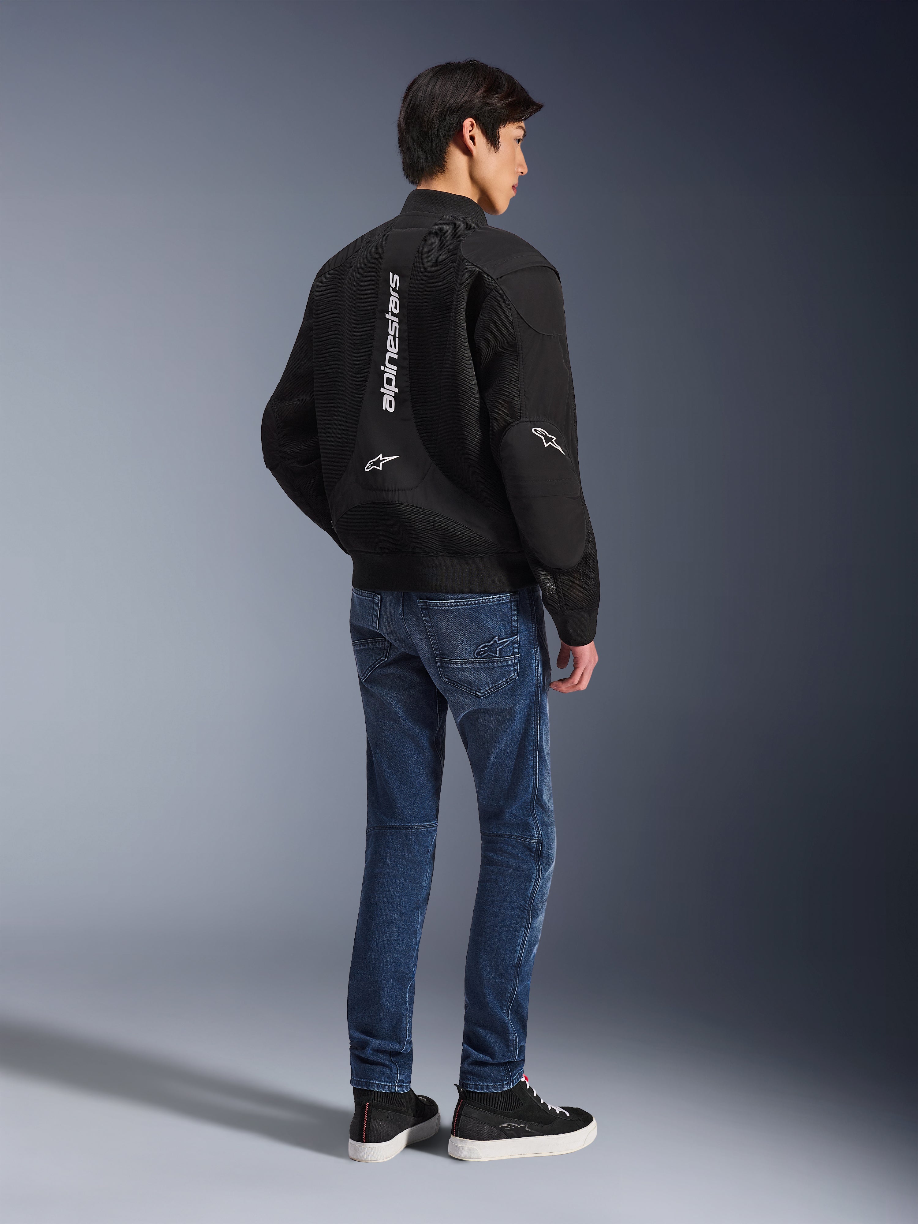 Model wearing Alpinestars Flight Air Jacket in Black, viewed from the back, paired with blue riding jeans and black sneakers, featuring vertical white Alpinestars branding and 3D mesh ventilation panels.