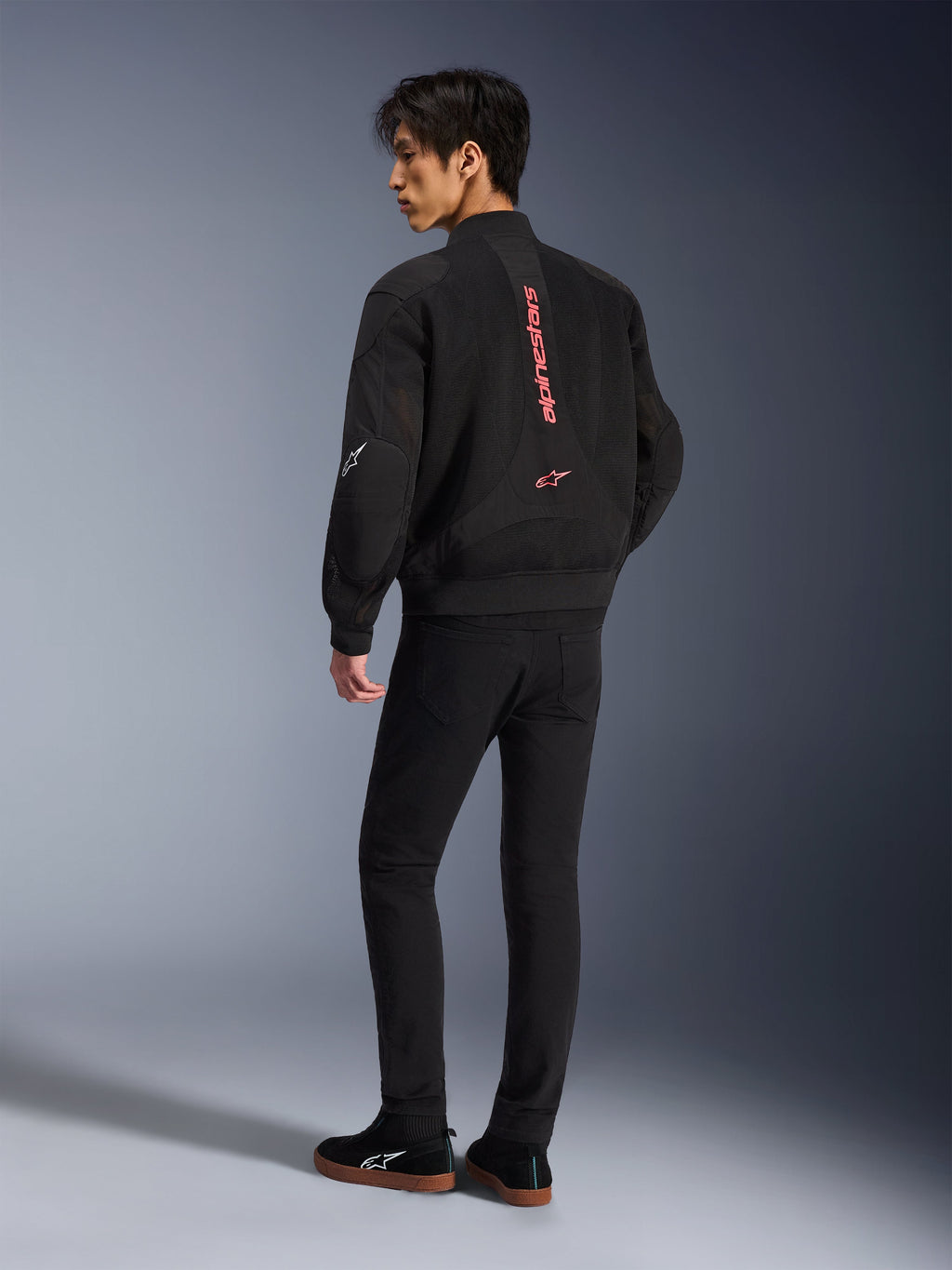Männliches Model trägt die Alpinestars Flight Air Jacke in Black Bright Red, Rückansicht mit vertikalem rotem Branding auf dem Rücken und 3D-Mesh-Konstruktion, kombiniert mit schwarzer Hose und Sneakern, urbaner Motorrad-Stil
