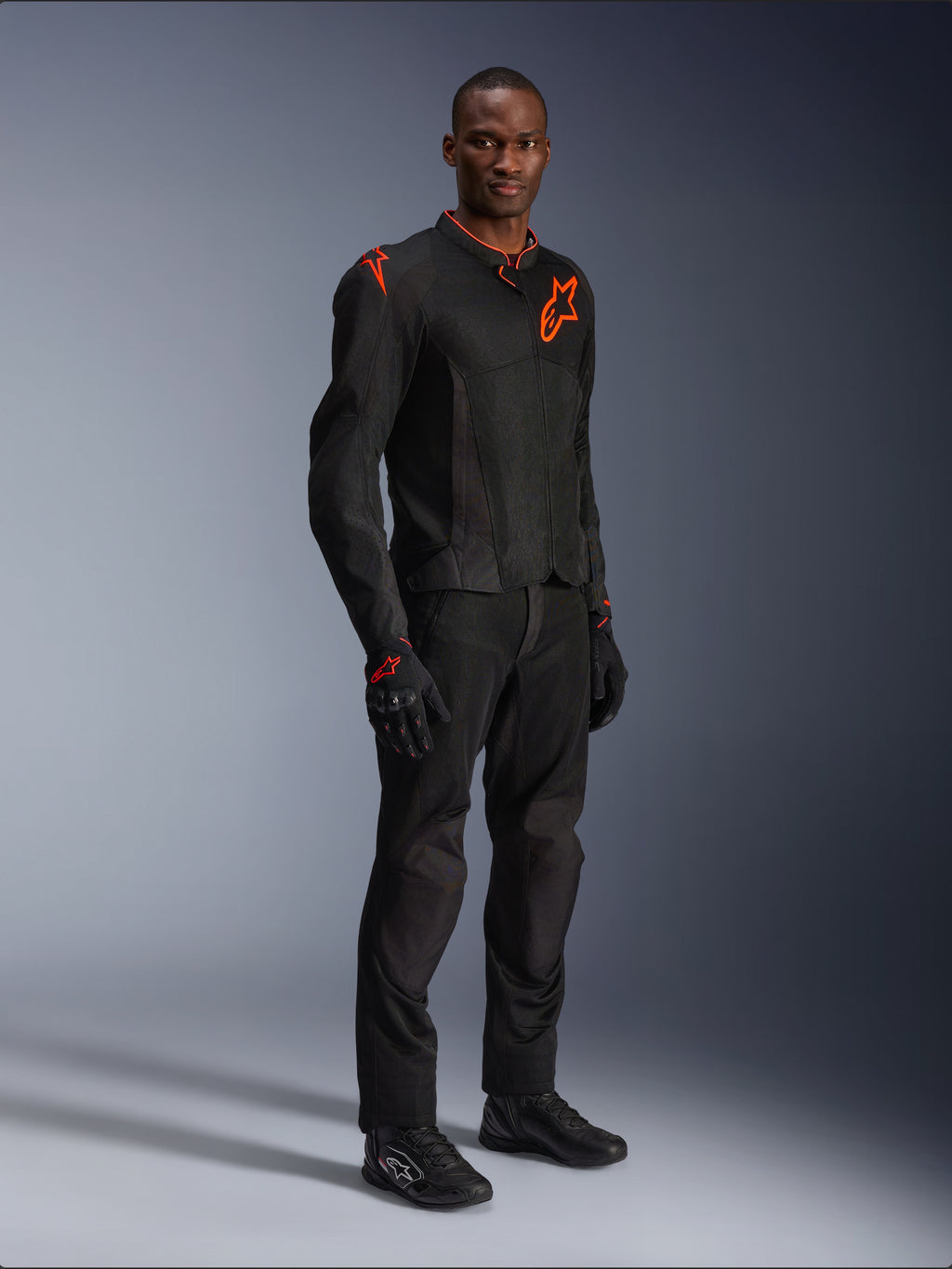Model in der Alpinestars Viper Air V4 Jacke in Schwarz und Red Fluo, mit Mesh-Einsätzen und roten Stern-Logos auf der Brust und Schulter, kombiniert mit passender schwarzer Motorradhose, Handschuhen und Fahrschuhen, professionelles Motorrad-Outfit für die Straße