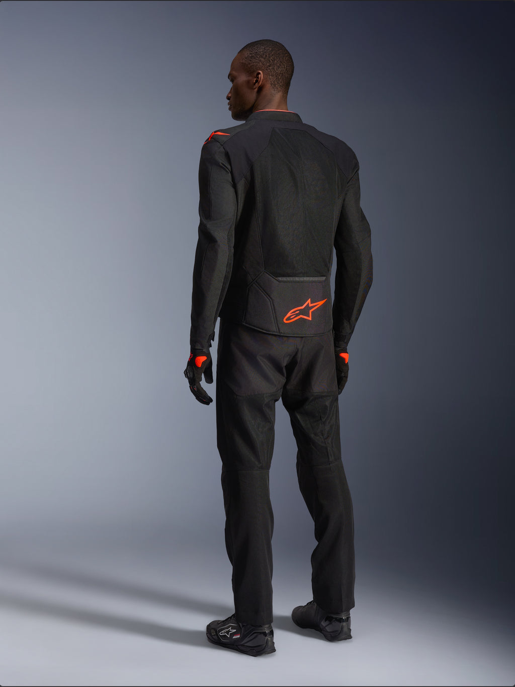 Männlicher Fahrer in der Rückansicht trägt die Alpinestars Viper Air V4 Jacke, Schwarz und Rot Fluo, mit einem orangefarbenen Astars-Logo auf dem unteren Rücken und den Schultern, Mesh-Textilkonstruktion, kombiniert mit passender schwarzer Motorradhose und Stiefeln.