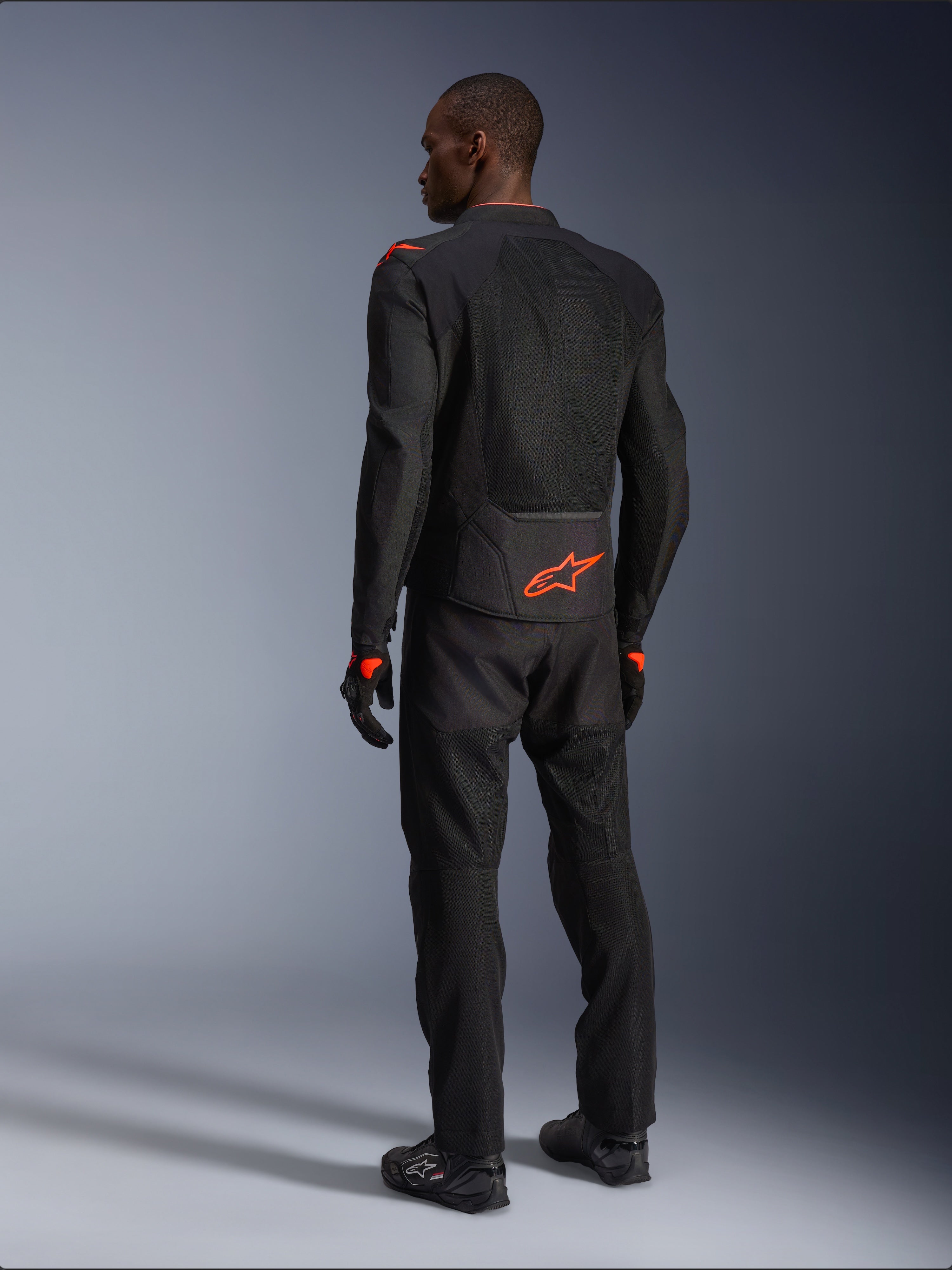 Männlicher Fahrer in der Rückansicht trägt die Alpinestars Viper Air V4 Jacke, Schwarz und Rot Fluo, mit einem orangefarbenen Astars-Logo auf dem unteren Rücken und den Schultern, Mesh-Textilkonstruktion, kombiniert mit passender schwarzer Motorradhose und Stiefeln.