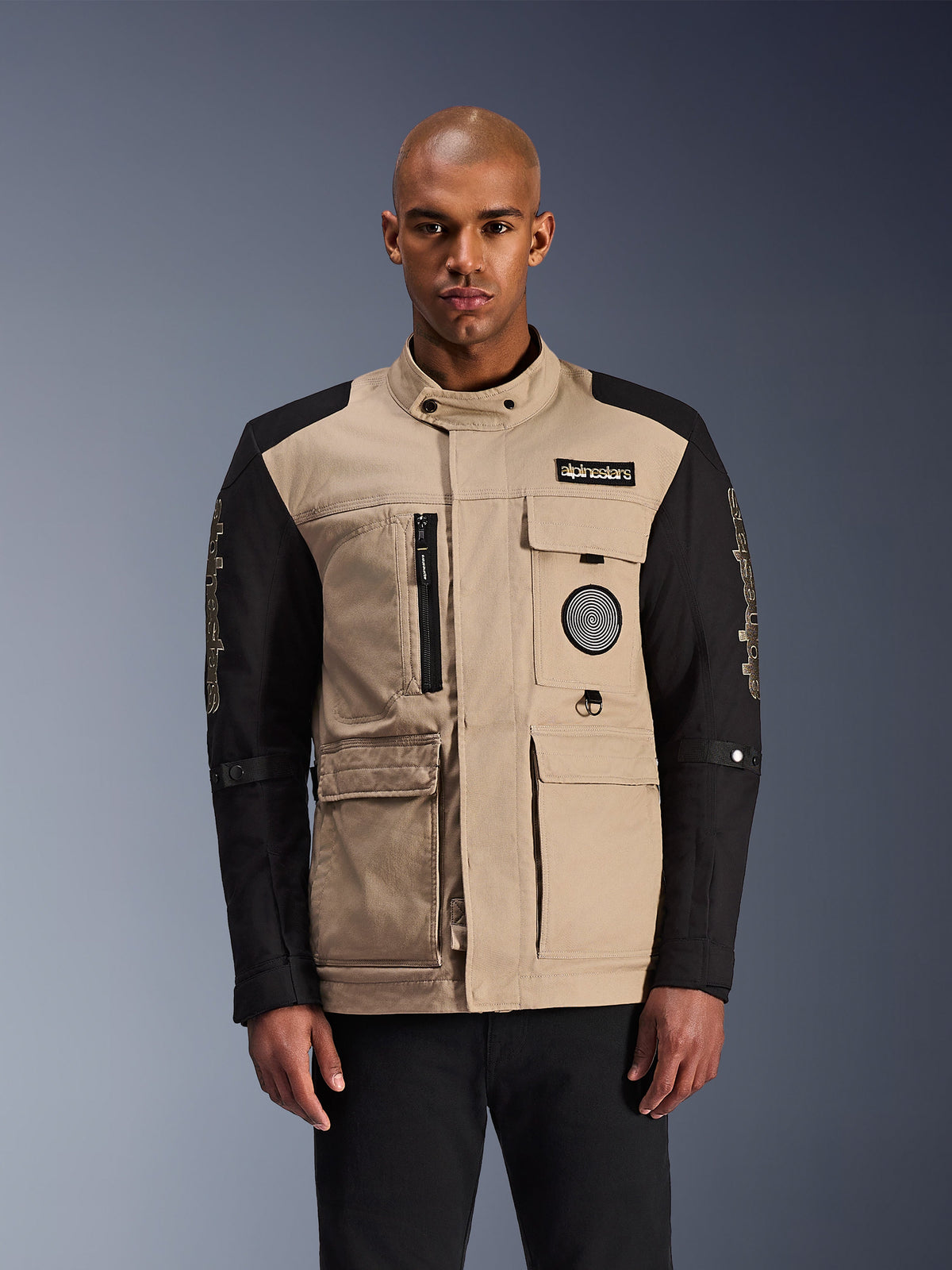 Alpinestars Dice Jacket, Urban Motorcycle Jacket, Beige and Black, rugged workwear aesthetic featuring multiple utility pockets and chest patches, high collar with button closure, bold branding on sleeves, worn by a male model