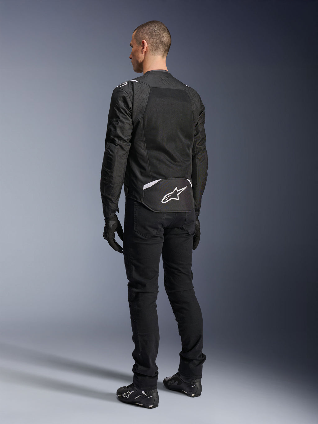 Alpinestars T-Sps Air V2 Jacke, Textil-Motorradjacke, Schwarz, Rückansicht mit großem Mesh-Belüftungspanel und weißem Alpinestars-Logo am unteren Rücken, kombiniert mit schwarzer Motorradhose und Handschuhen für einen sportlichen urbanen Look.