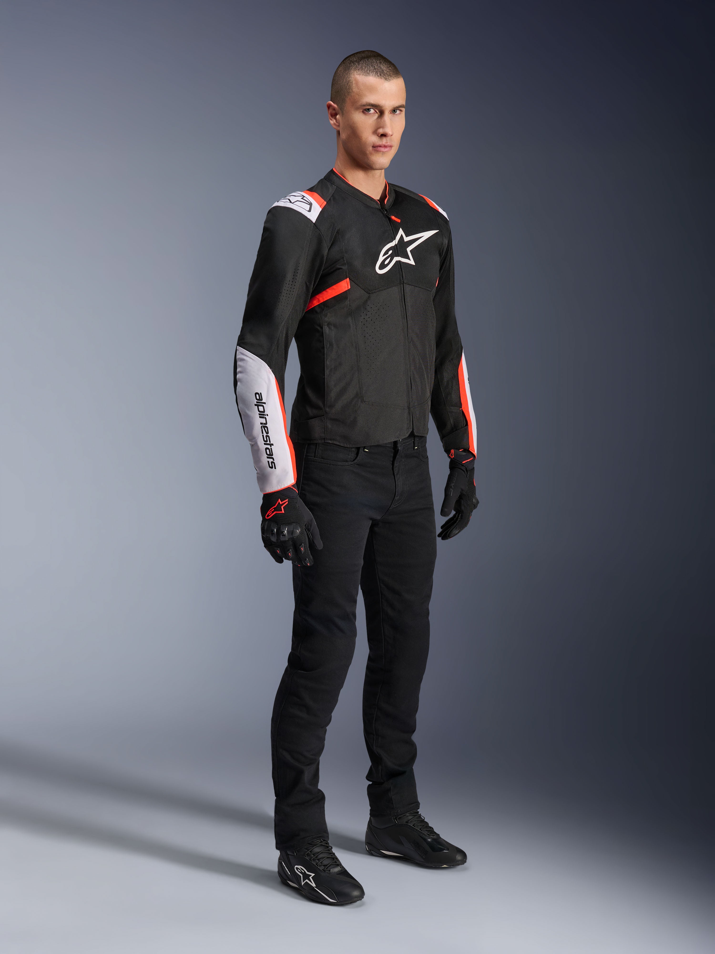 Alpinestars T-Sps Air V2 Jacke, Schwarz Weiß Rot Fluo, getragen von einem männlichen Model mit schwarzer Motorradhose und Handschuhen, Textil-Motorradjacke mit großen Mesh-Einsätzen, weißen Schulter- und Unterarmbereichen, markantes Alpinestars-Logo auf der Brust.