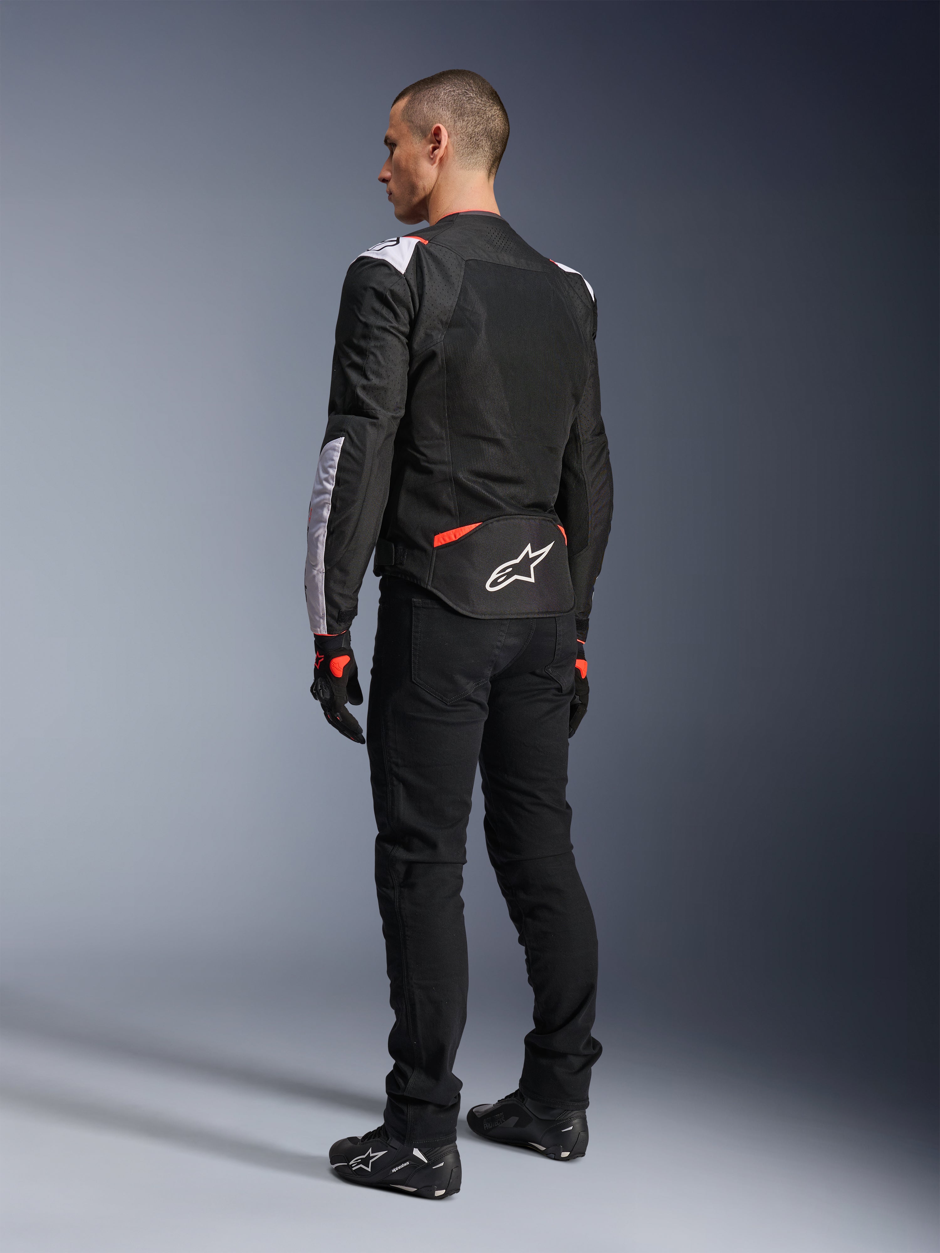 Alpinestars T-Sps Air V2 Jacke, Textil-Motorradjacke, Schwarz, Weiß und Fluo-Rot, Rückansicht am Modell, Mesh-Einsätze für Belüftung, weißes Astars-Logo am unteren Rücken, kombiniert mit schwarzer Motorradhose und Handschuhen