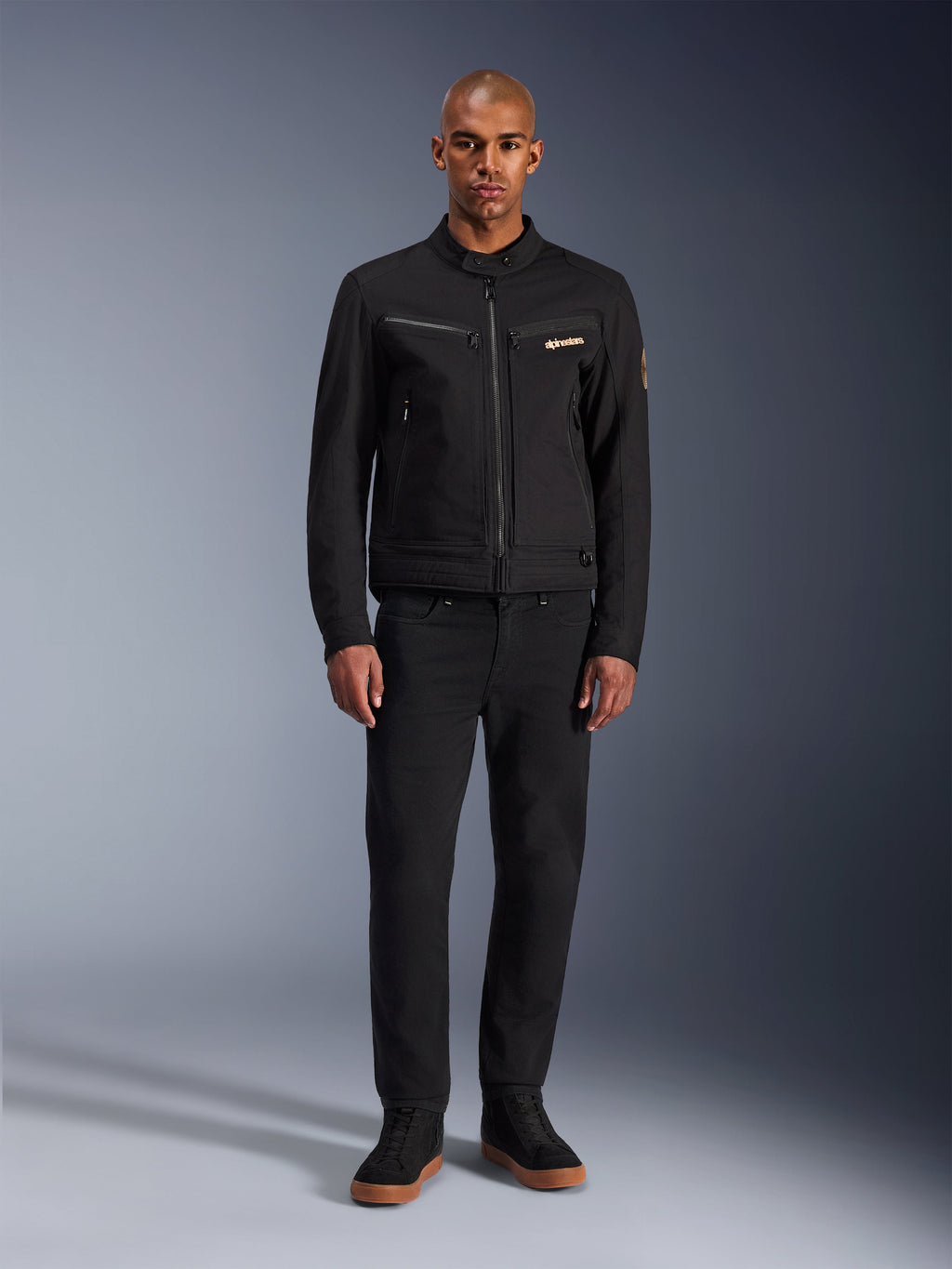 Alpinestars Bruiser Jacke, schwarze urbane Motorradjacke, getragen von einem Model mit schwarzer Hose und High-Top-Sneakern, Vorderansicht mit Brusttaschen mit Reißverschluss und besticktem Logo, klassisches, von Workwear inspiriertes Design für den täglichen Weg zur Arbeit