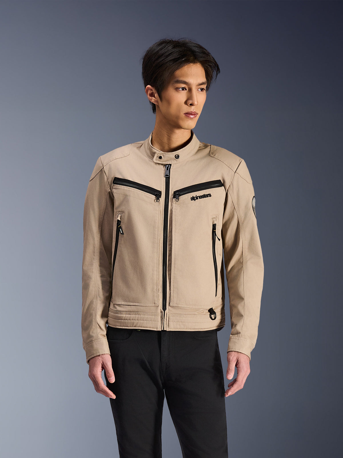 Alpinestars Bruiser Jacke, beige Motorradjacke getragen von einem männlichen Model, schwarze Reißverschlussdetails an Brust- und Seitentaschen, hoher Druckknopfkragen, Workwear-inspirierte urbane Motorradbekleidung, kombiniert mit einer schwarzen Hose