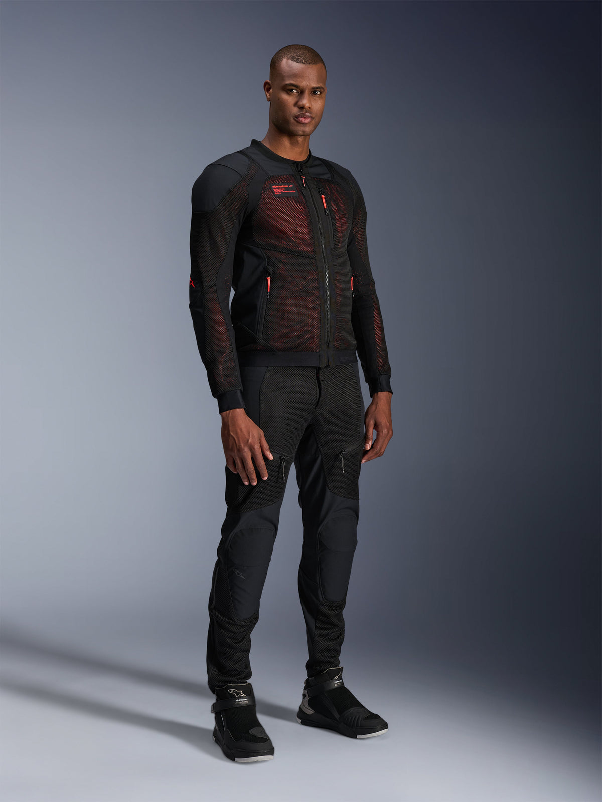 Alpinestars Troop-Air Pro Jacke in Schwarz Rot Fluo, technische gepanzerte Mesh-Motorradjacke, getragen von einem männlichen Model mit passender schwarzer gepanzerter Hose und High-Top-Stiefeln, Ganzkörper-Studioporträt.