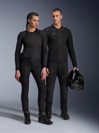 Alpinestars Stella Troop-Air Jacke, Mann und Frau tragen passende, komplett schwarze technische Motorradausrüstung, mit Mesh-Konstruktion für optimale Belüftung, schwarze Hosen und Stiefel, der Mann hält einen schwarzen Helm, urbane Motorradbekleidung