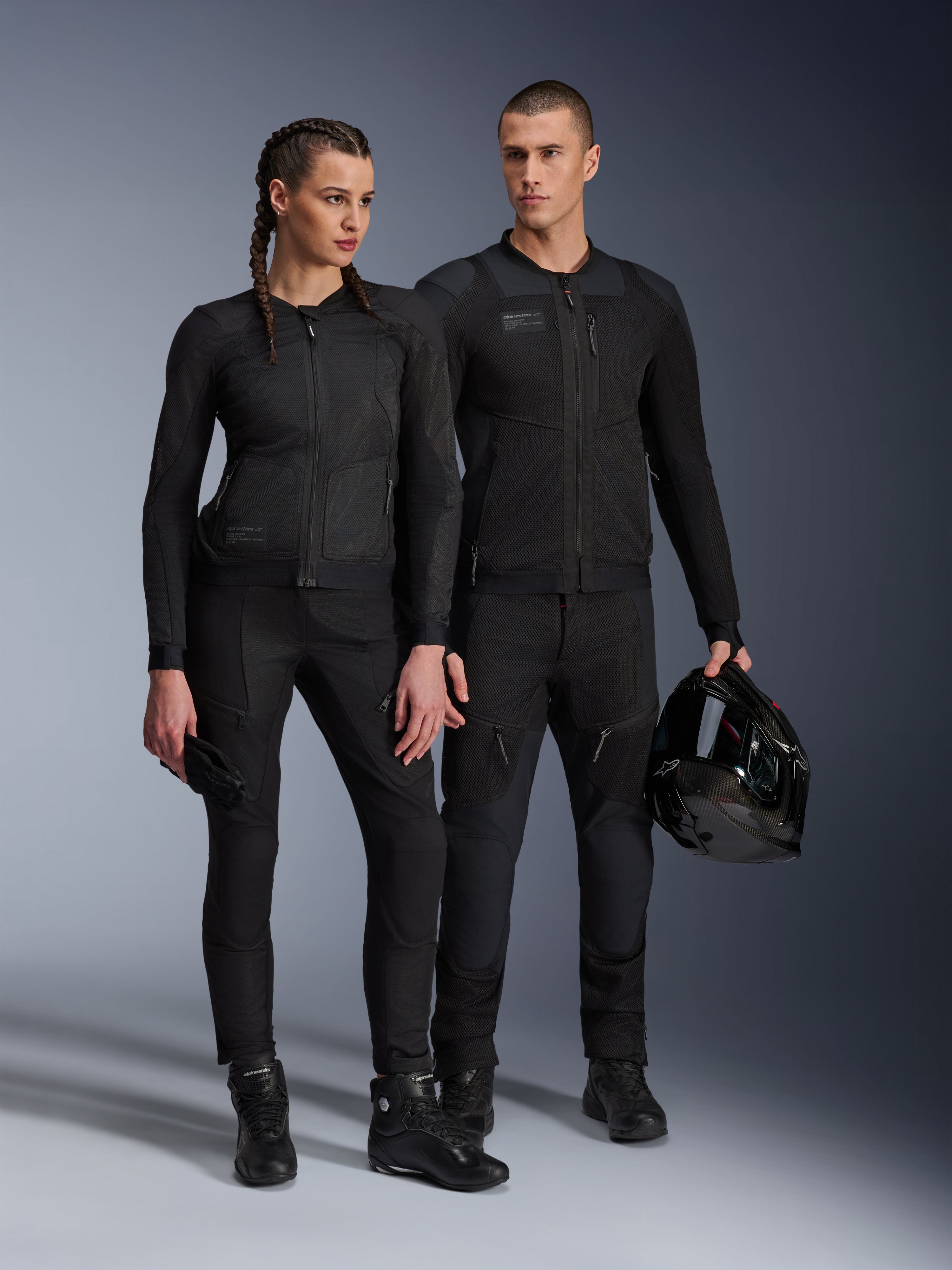 Alpinestars Stella Troop-Air Jacke, Mann und Frau tragen passende, komplett schwarze technische Motorradausrüstung, mit Mesh-Konstruktion für optimale Belüftung, schwarze Hosen und Stiefel, der Mann hält einen schwarzen Helm, urbane Motorradbekleidung