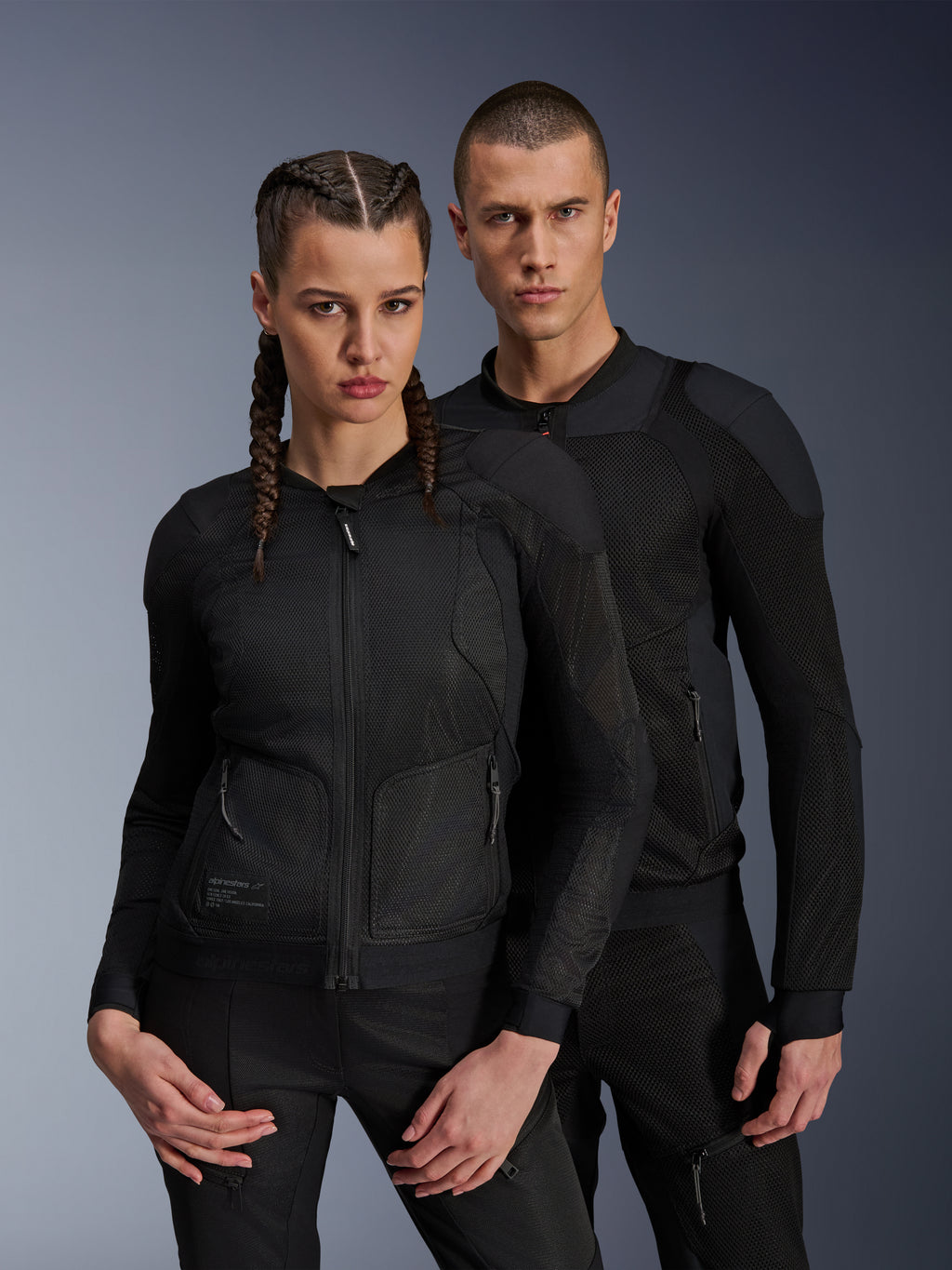 Mann und Frau in Alpinestars Troop-Air und Stella Troop-Air Jacken, schwarze technische Mesh-Motorradjacken mit 2D-Tech-Mesh-Struktur zur Belüftung und verstärkten Einsätzen, moderne urbane Fahrbekleidung.