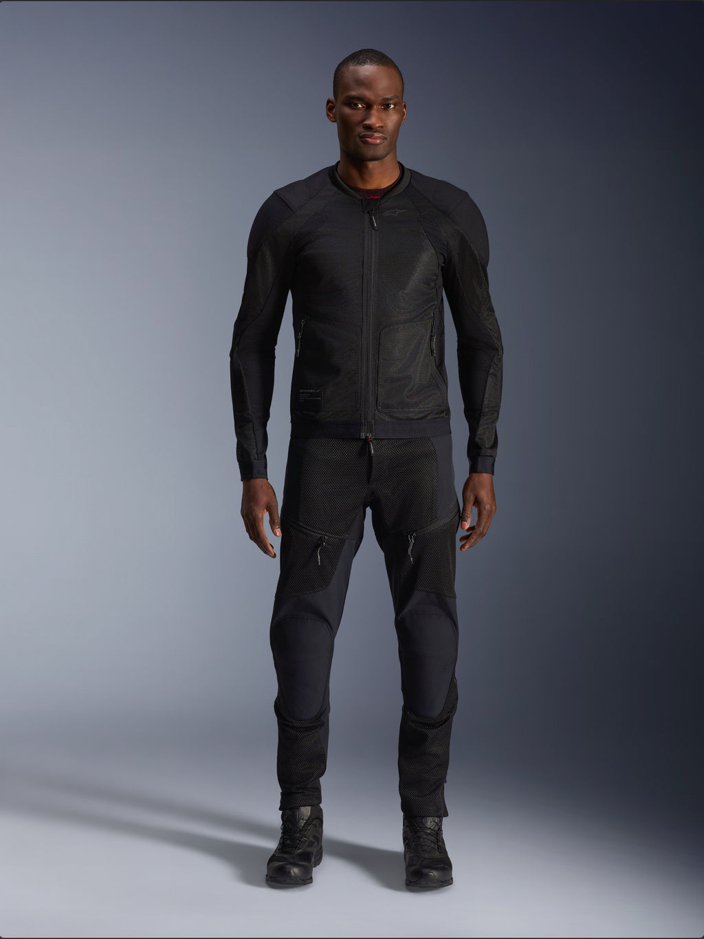 Männliches Model in der Alpinestars Troop-Air Jacke in Black Black, leichte Mesh-Motorradjacke, kombiniert mit passender schwarzer Motorradhose und Stiefeln, mit technischer 2D-Tech-Mesh-Konstruktion für eine überlegene Belüftung.